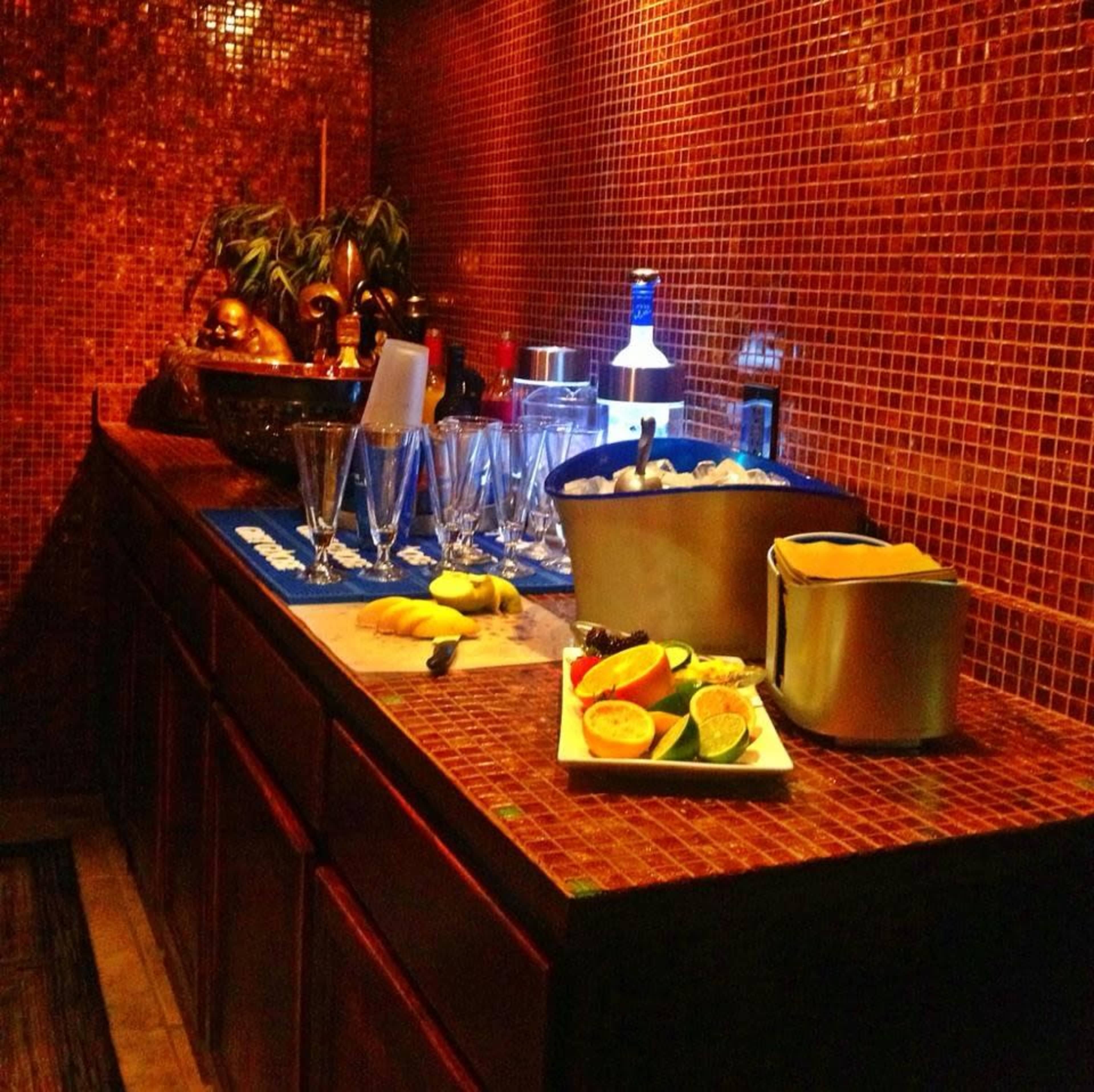 A bar station is set against a mosaic tile wall, featuring various drinks, garnishes, and glassware arranged on a countertop.