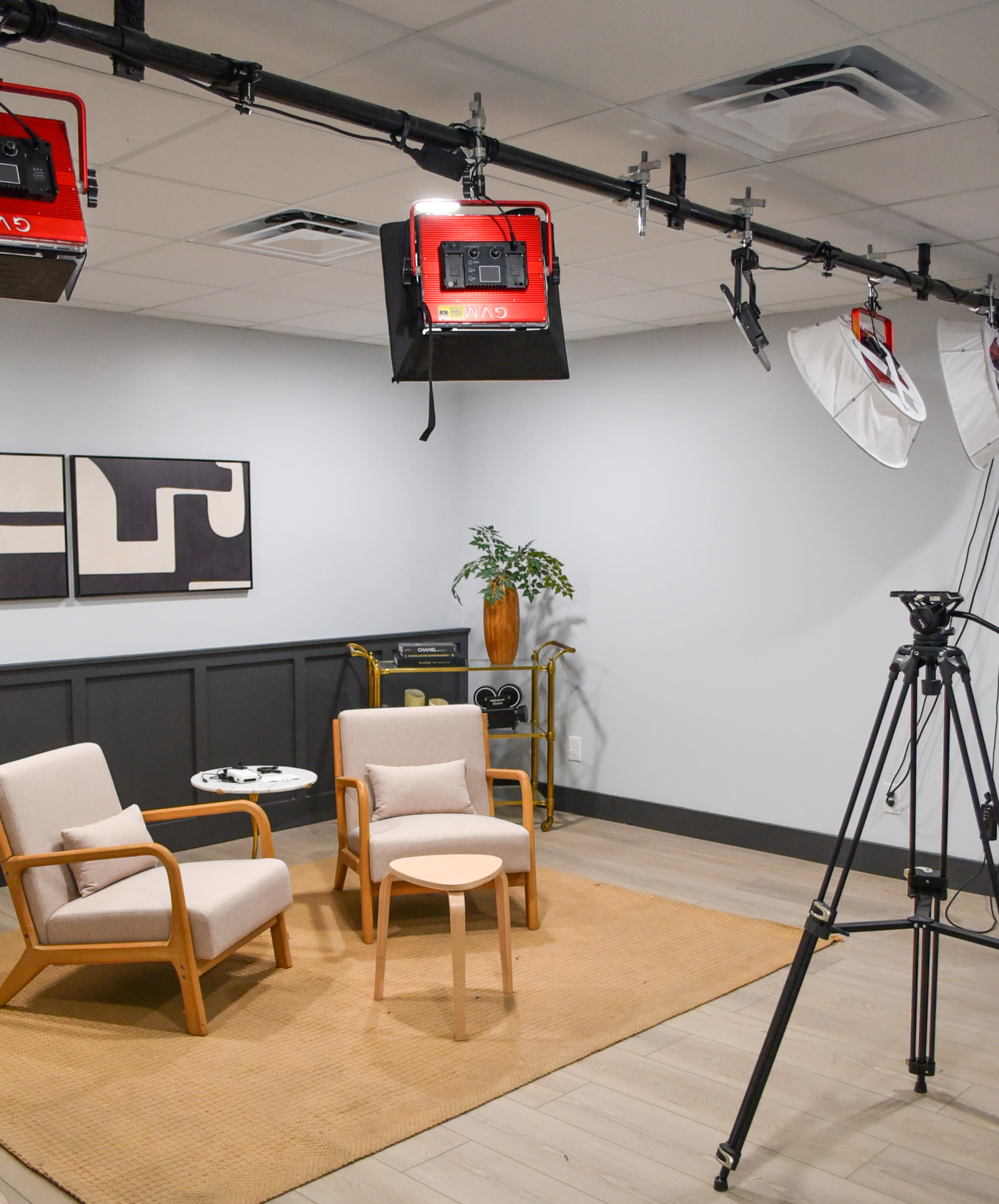 A well-lit studio setup features two wooden chairs and a small table on a rug, with lighting equipment and a camera on a tripod.
