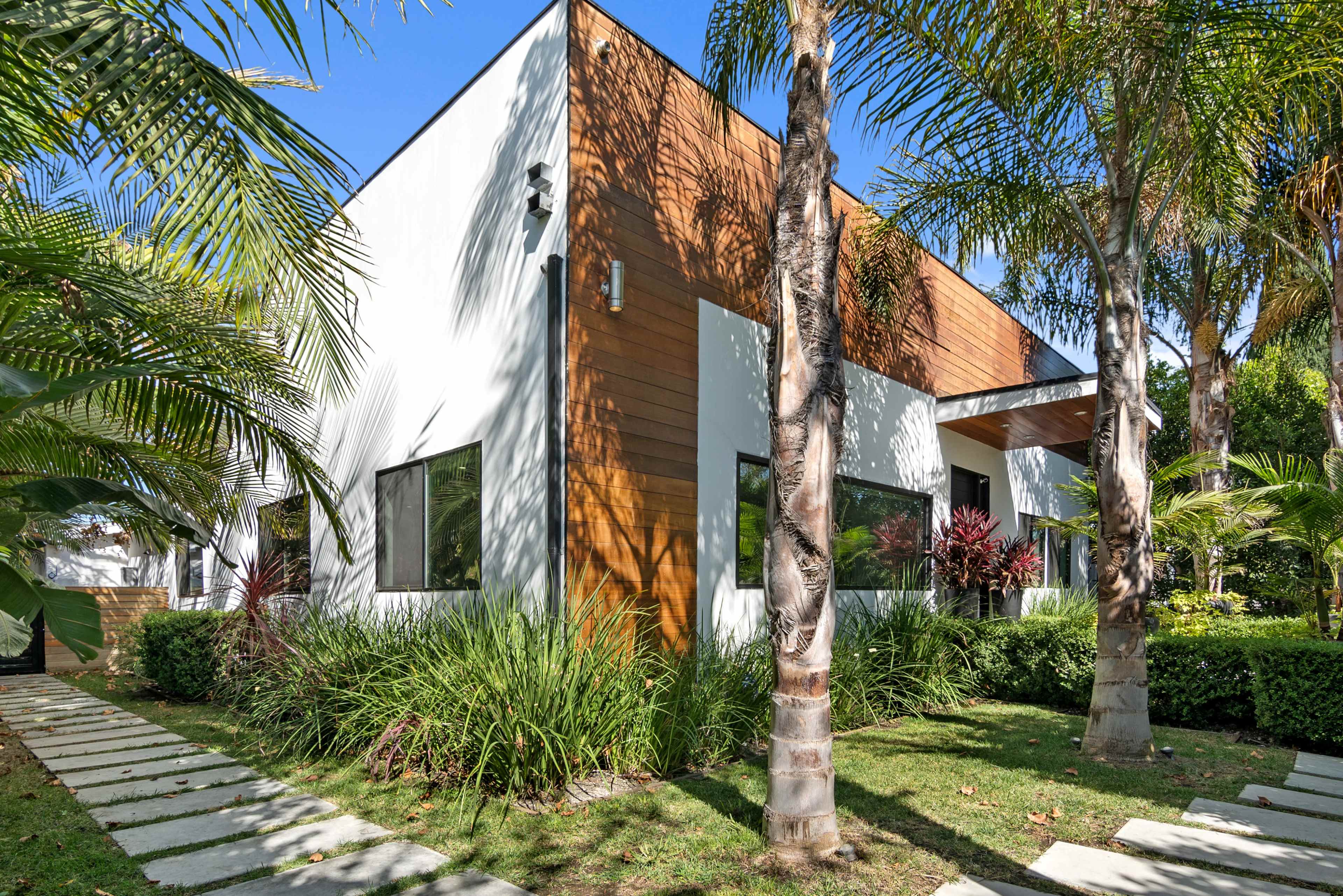 A modern house with a combination of white and wood paneling is surrounded by palm trees and landscaped greenery.