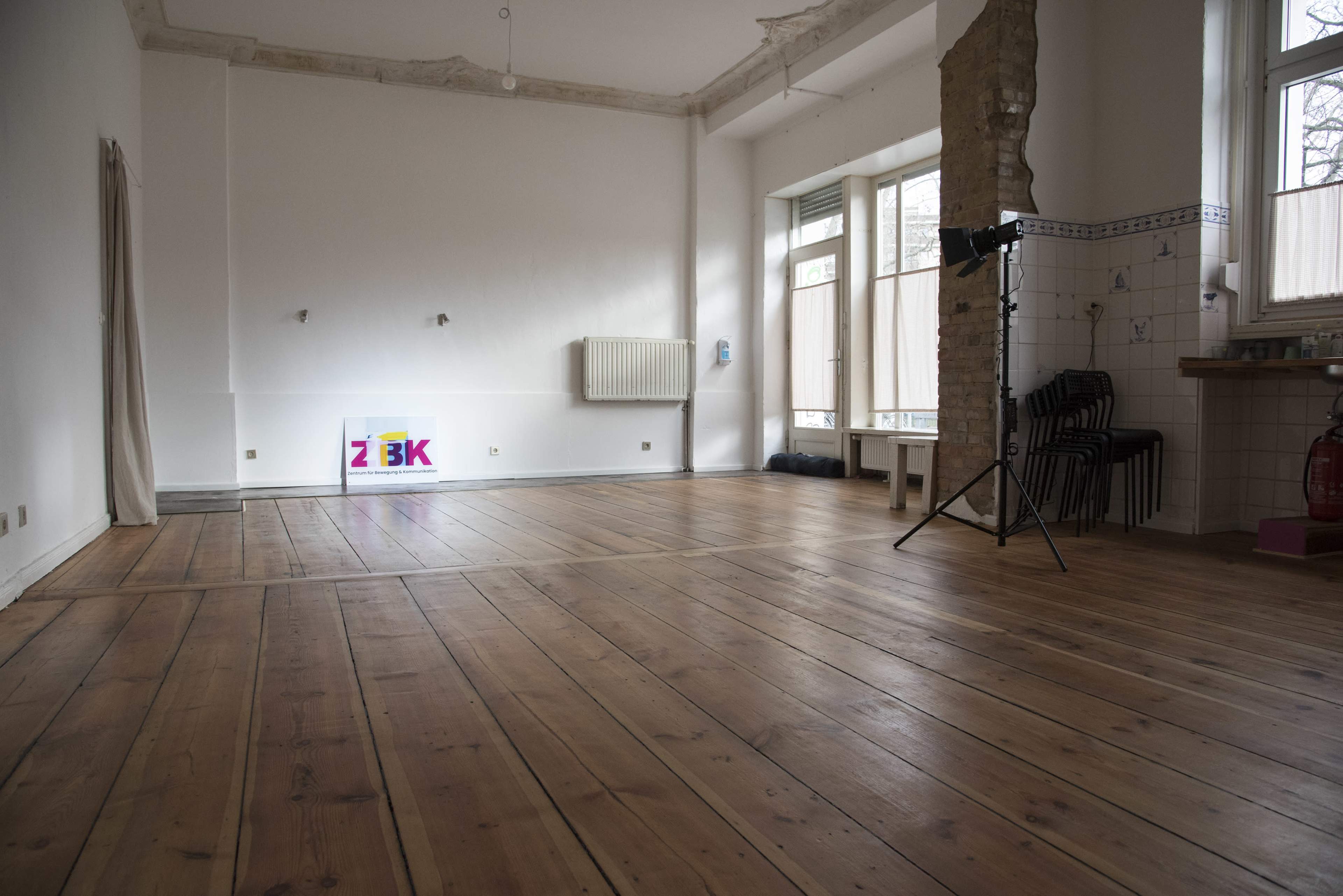 A spacious, empty room with wooden floors, a large window, and minimal furnishings including a radiator and a light stand.