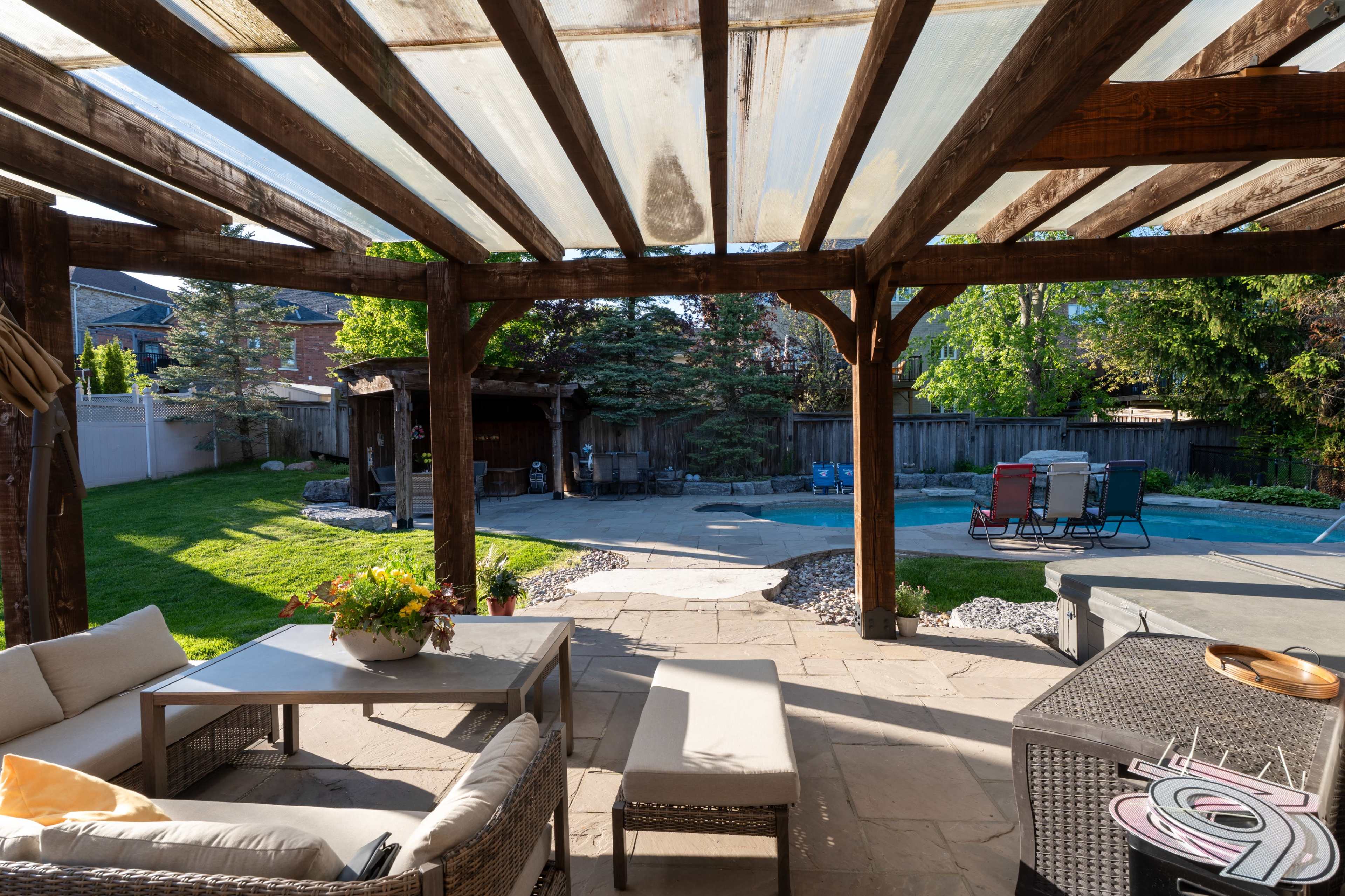 The image shows a backyard patio area with wooden beams overhead, furnished with a table and chairs, overlooking a lawn and a pool area.