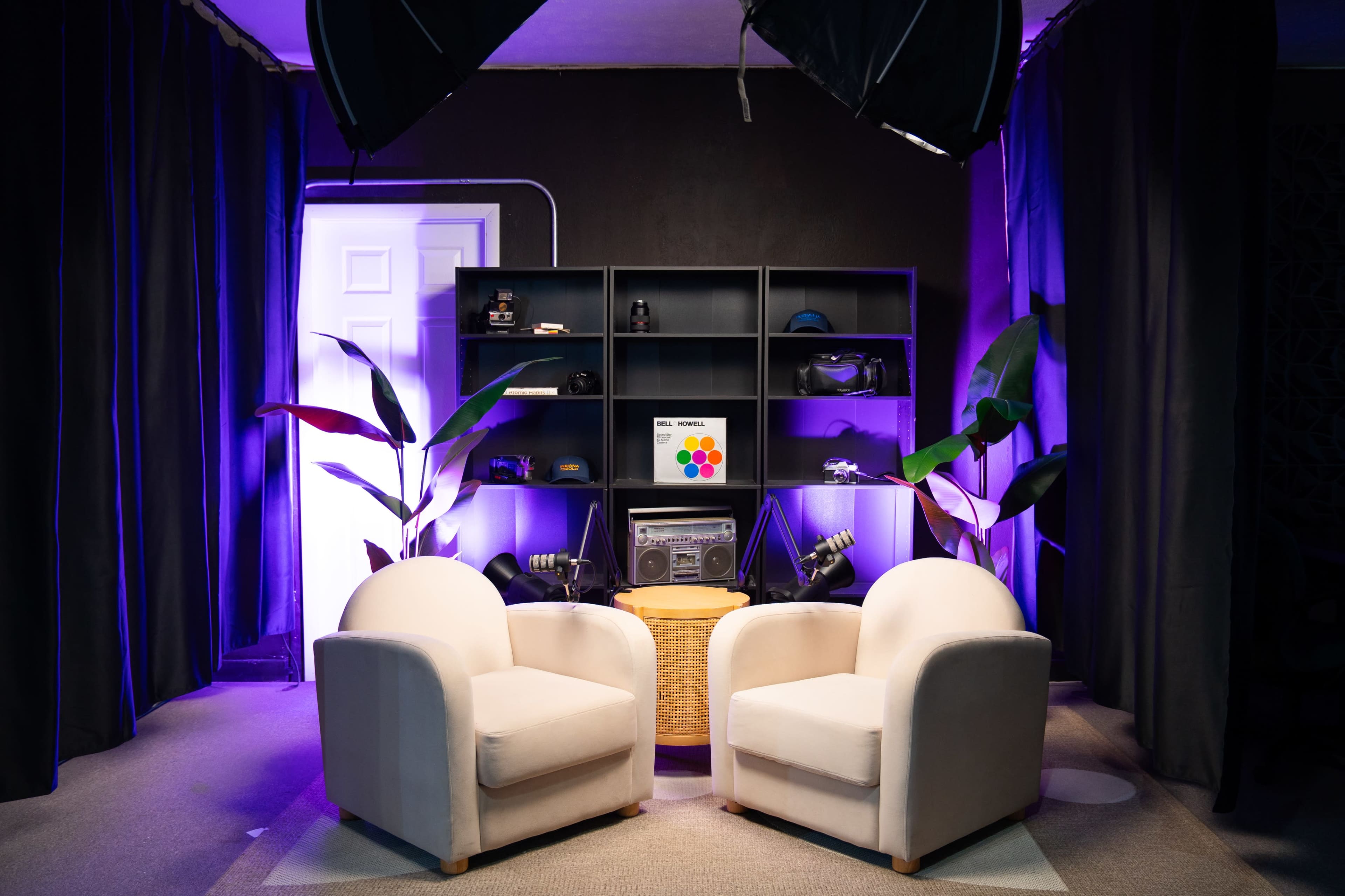 The image shows a cozy studio setup with two light-colored chairs facing each other in front of a shelf displaying various items, illuminated by purple lighting.