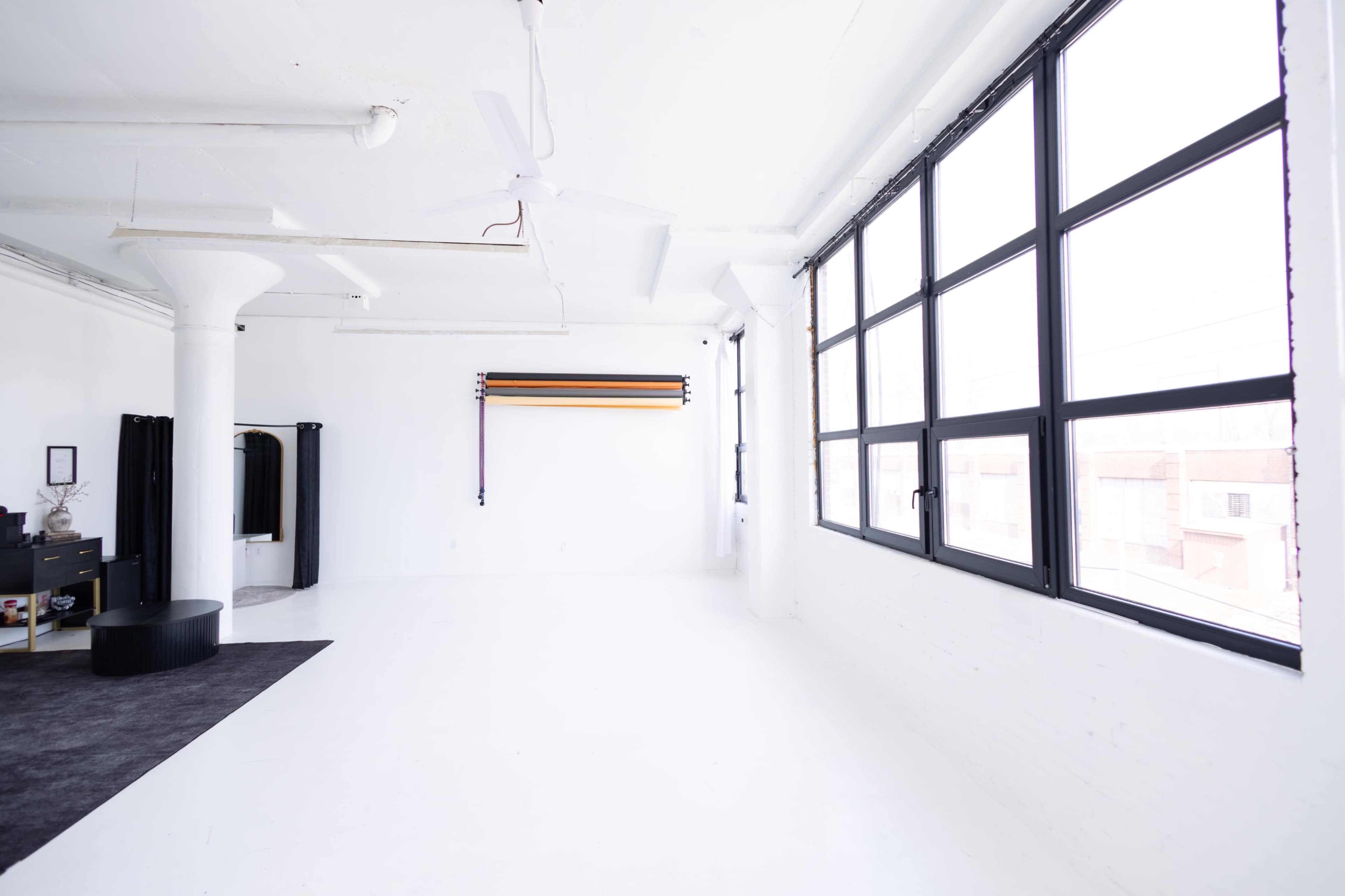 The image shows a bright, open studio space with white walls, large windows, and minimal furniture.