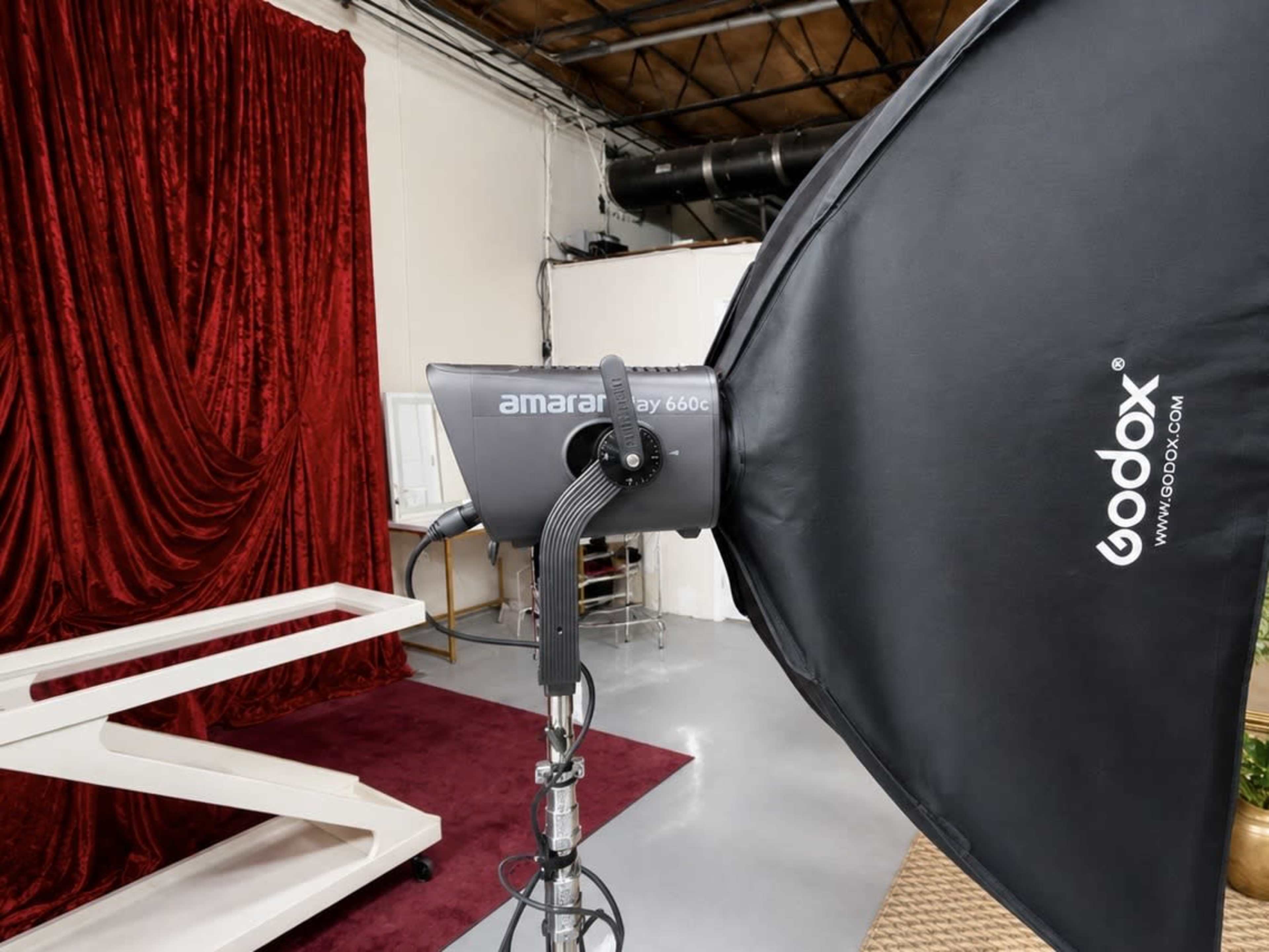 The image shows a photography lighting equipment setup with a Godox Amaran light mounted on a stand, positioned in front of a red velvet backdrop.