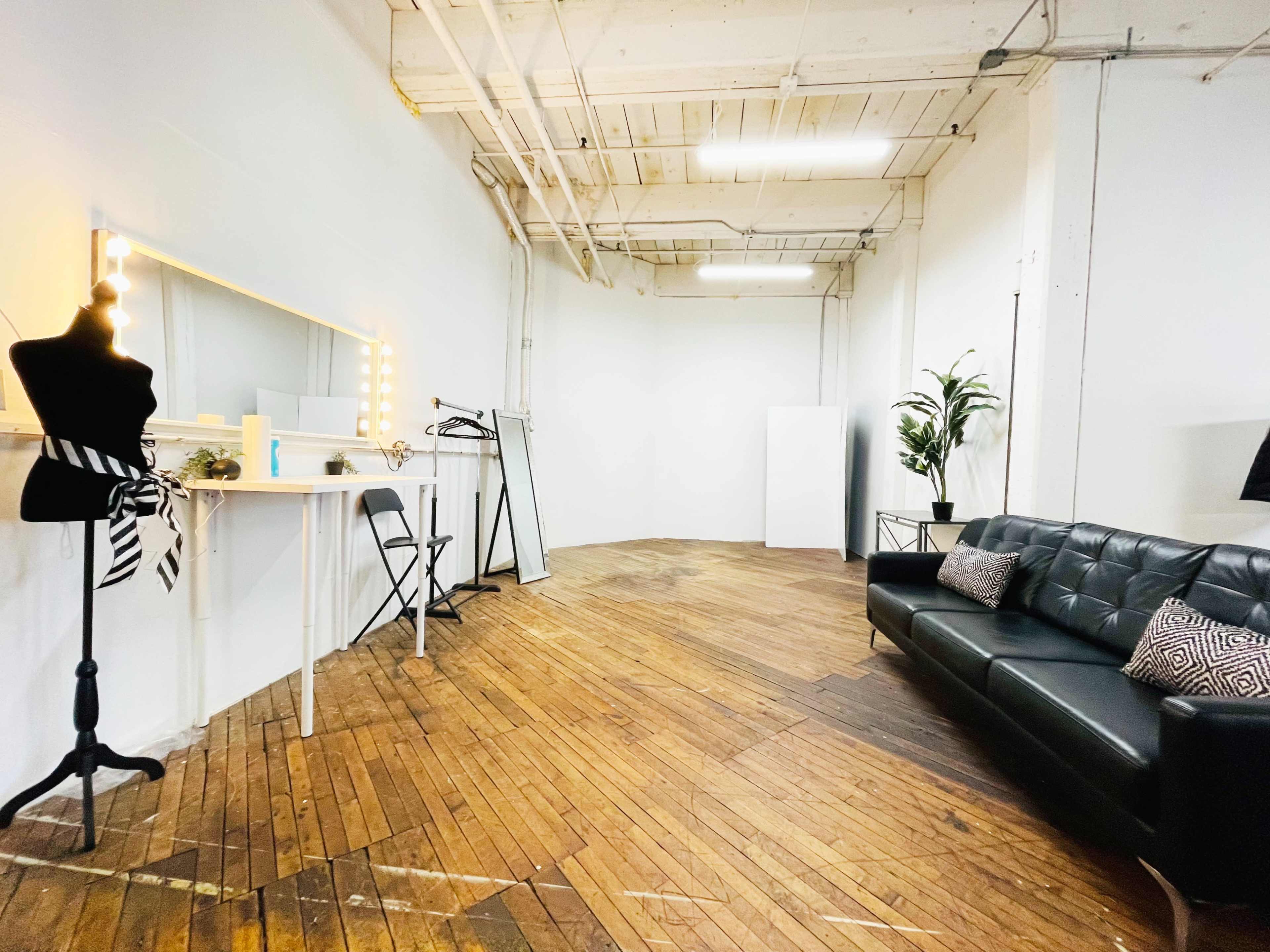 A spacious, well-lit room features a black leather sofa, a mannequin, and a large mirror surrounded by lights on a wooden floor.