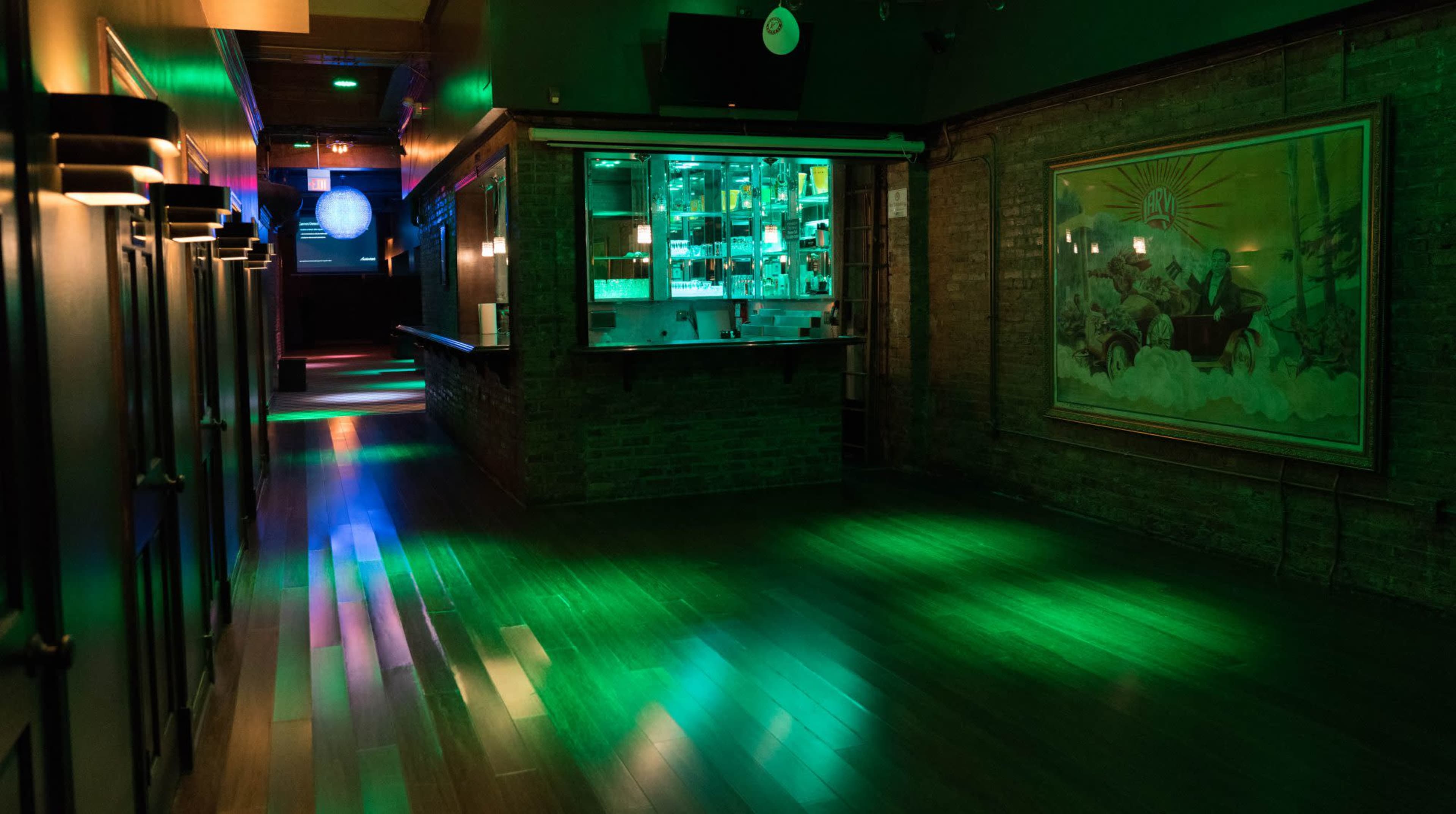 The image shows a dimly lit bar area with colorful lights reflecting on a wooden floor and a large framed painting on the wall.