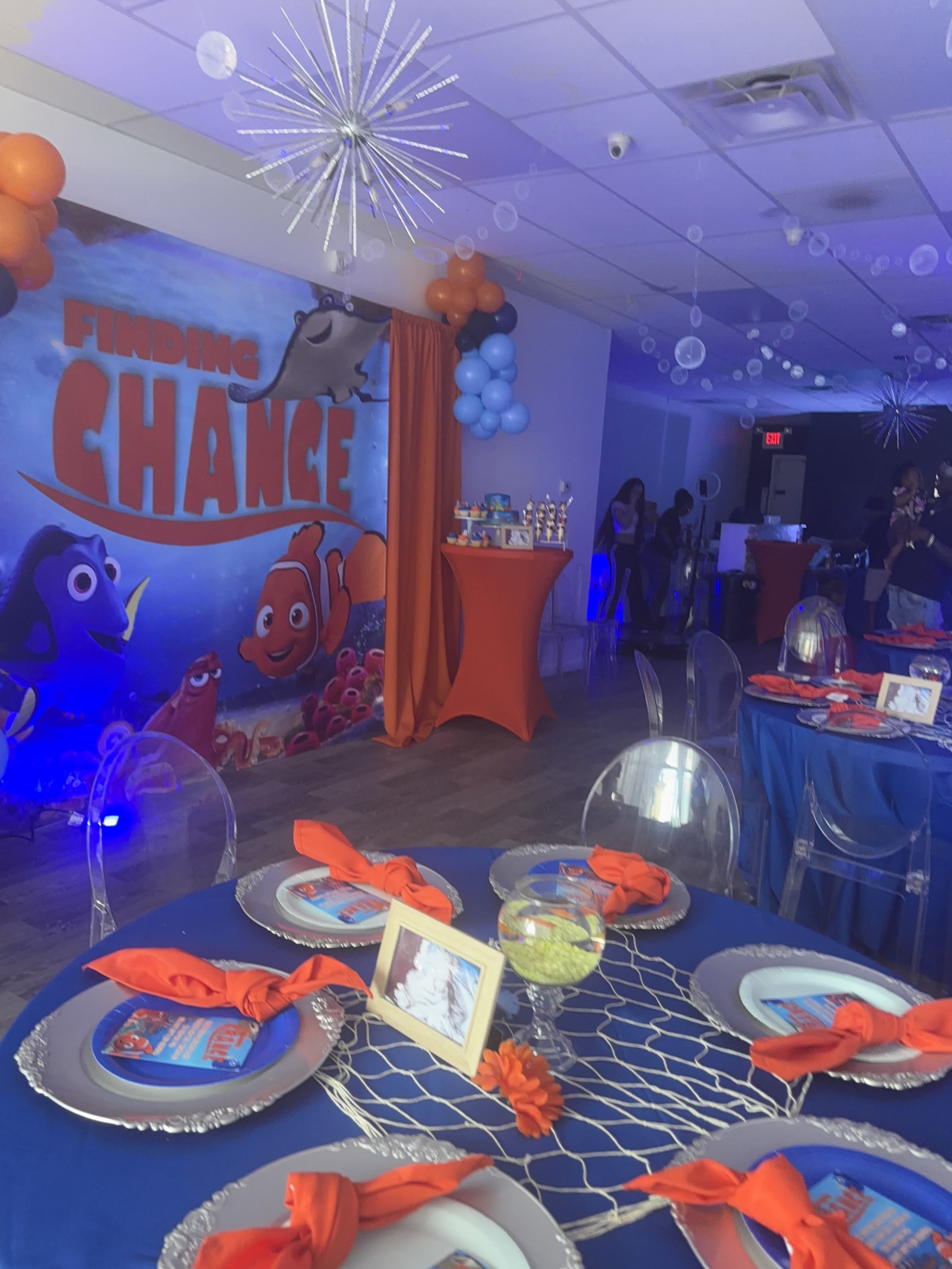 The image shows a decorated party space featuring a "Finding Change" theme, complete with colorful balloons, tables set with orange napkins, and a backdrop with aquatic characters.