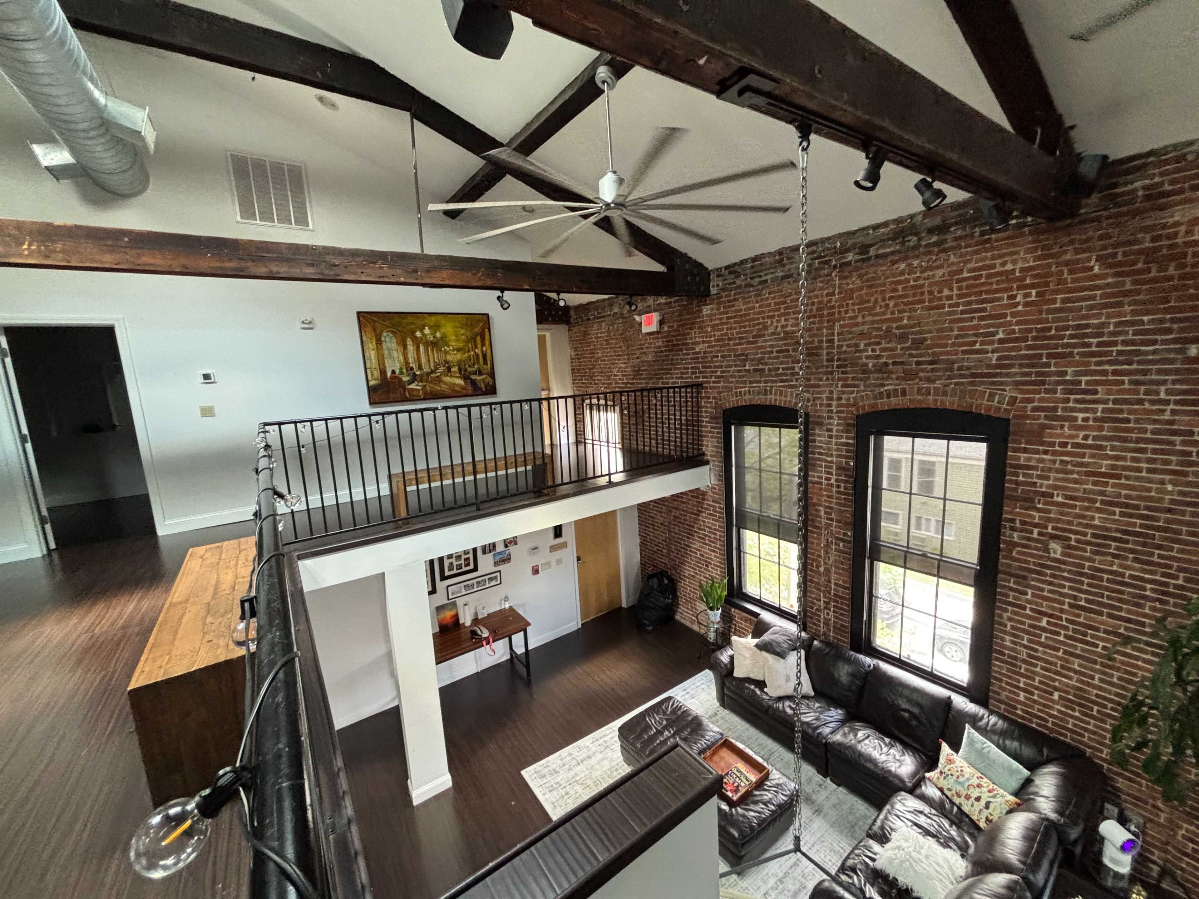 The image shows a spacious loft-style living area with exposed brick walls, large windows, and a second-level balcony overlooking a seating area featuring a sectional couch.