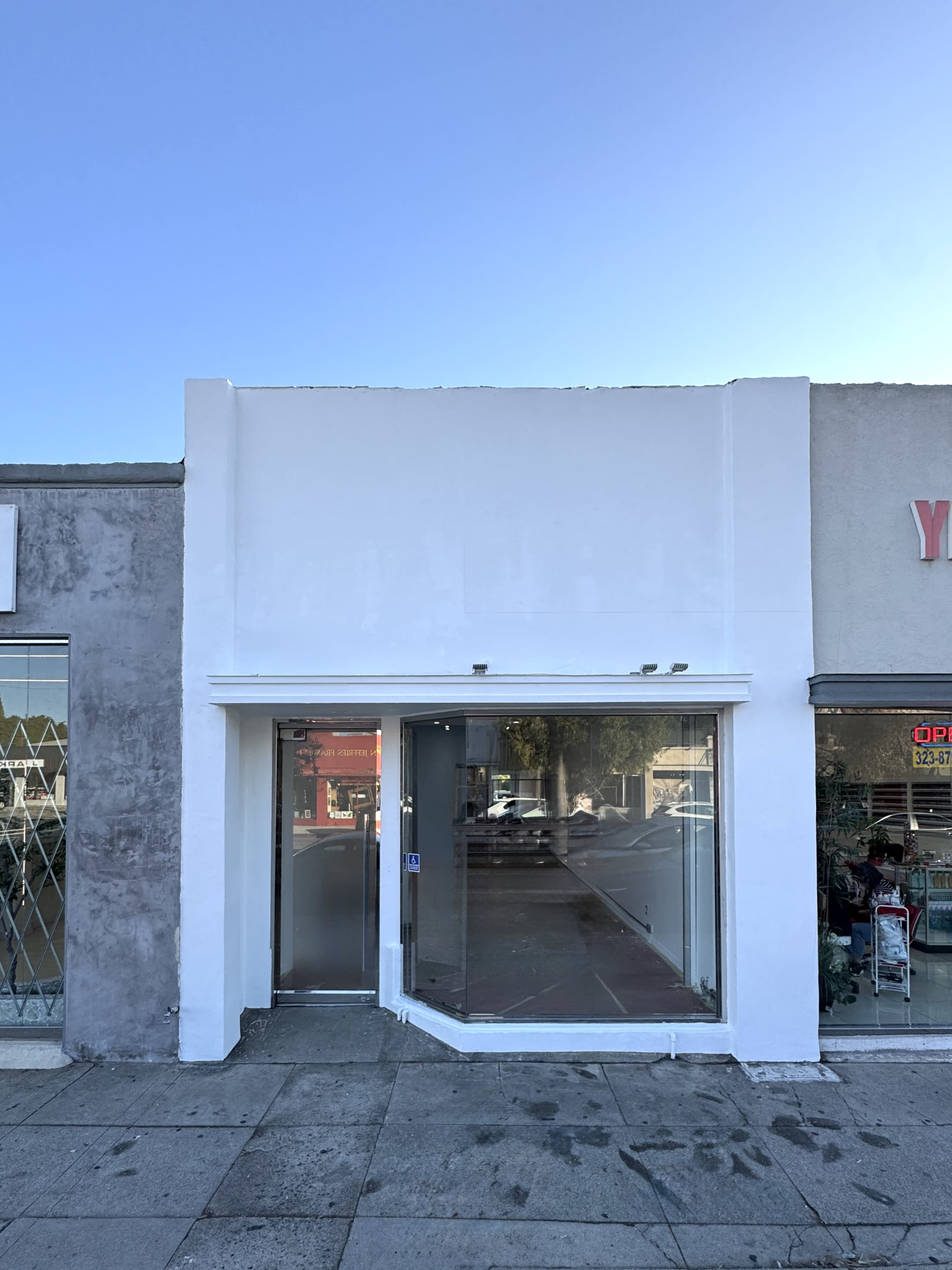 The image shows a vacant storefront with a large glass window and a white facade, situated between two neighboring businesses.