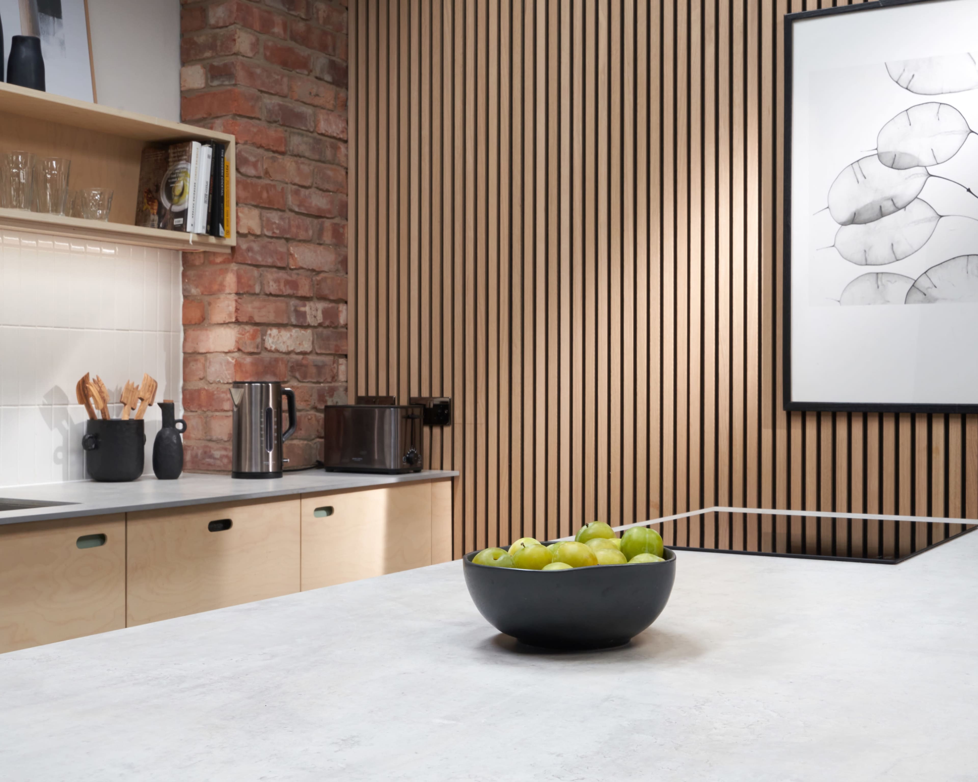 A modern kitchen featuring a wooden accent wall, a concrete countertop, and a black bowl of green apples on a central island.