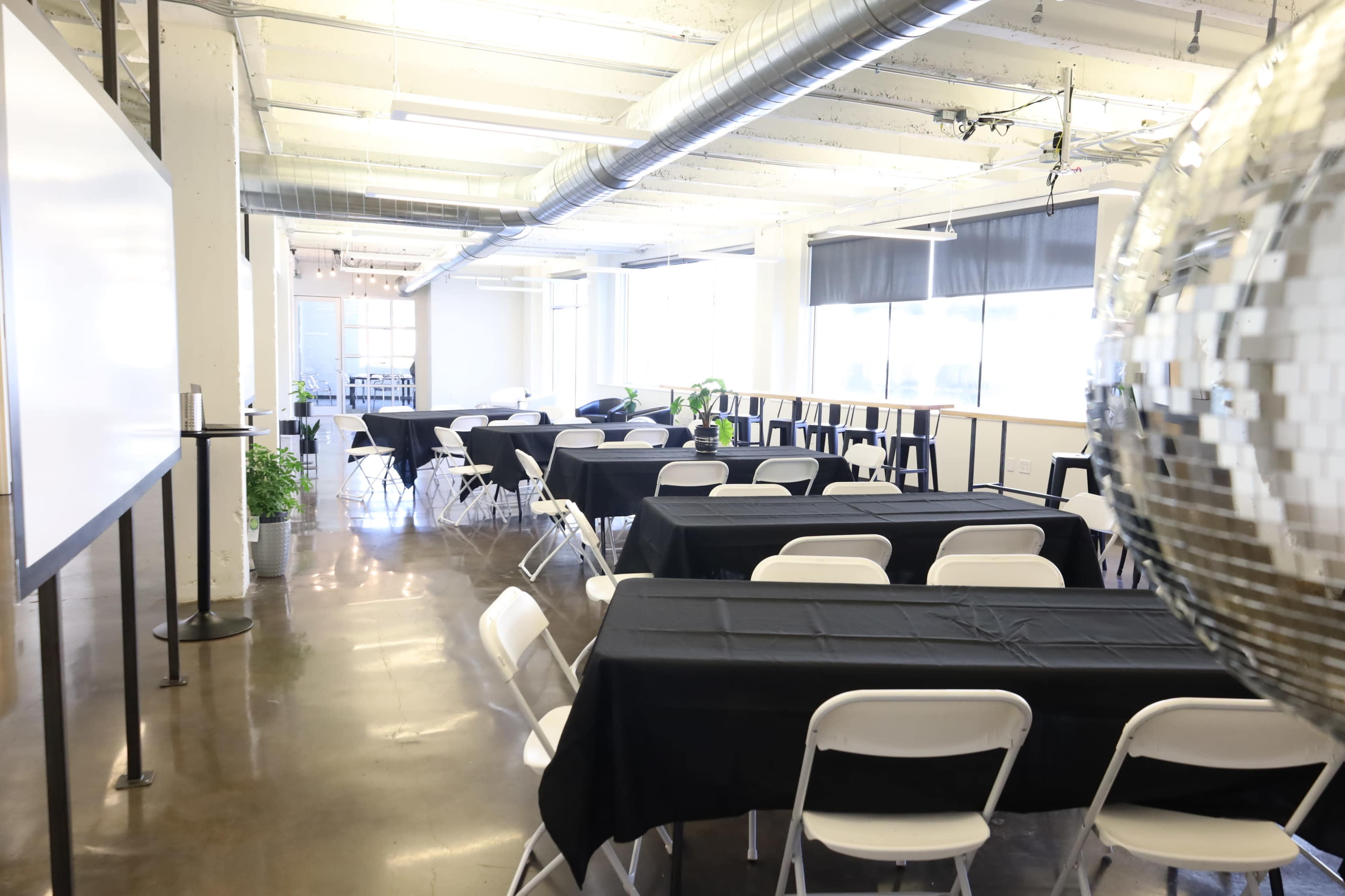 The image shows a spacious event room set up with several long tables covered in black cloth and white chairs arranged throughout the space.