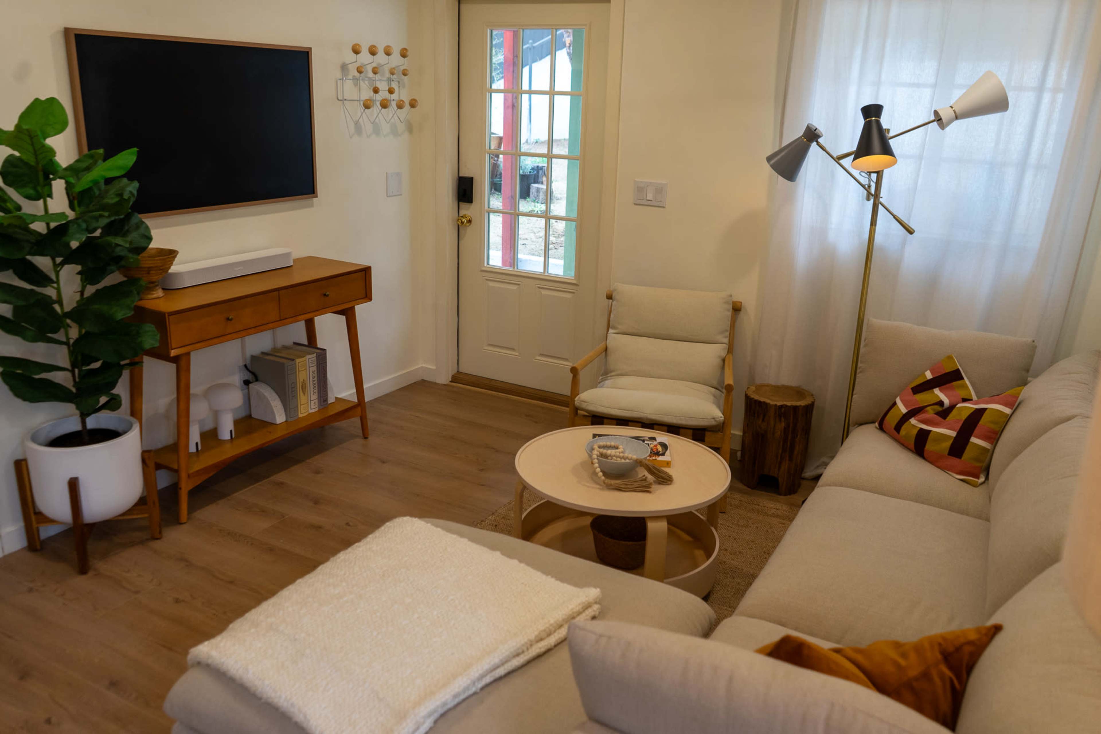 The image shows a cozy living room with a beige sectional sofa, a round coffee table, a wall-mounted television, and a door leading outside, complemented by indoor plants and varying furniture pieces.