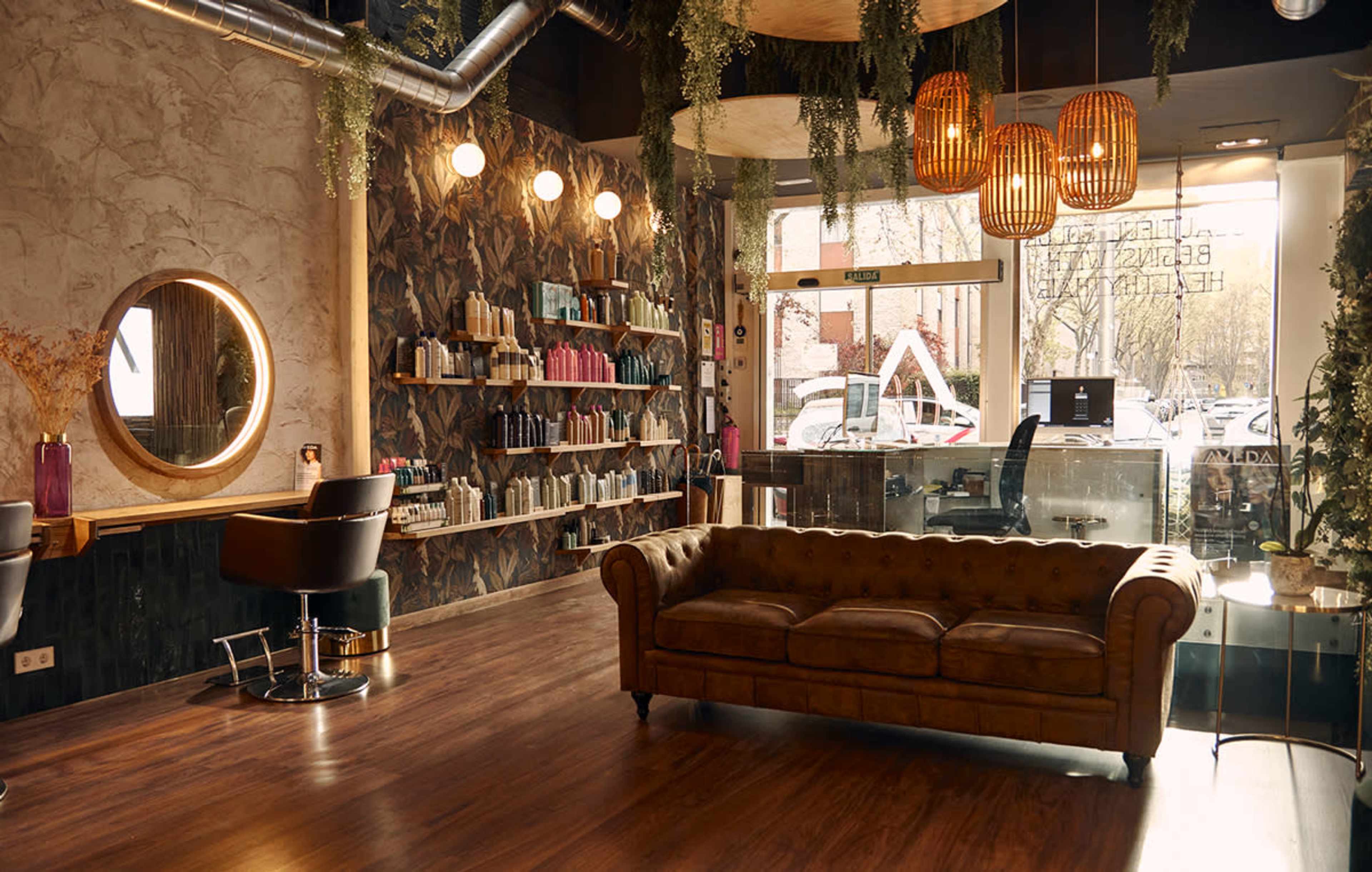 The image features a stylish hair salon with a brown leather couch, wooden shelves filled with hair products, and decorative light fixtures hanging from the ceiling.