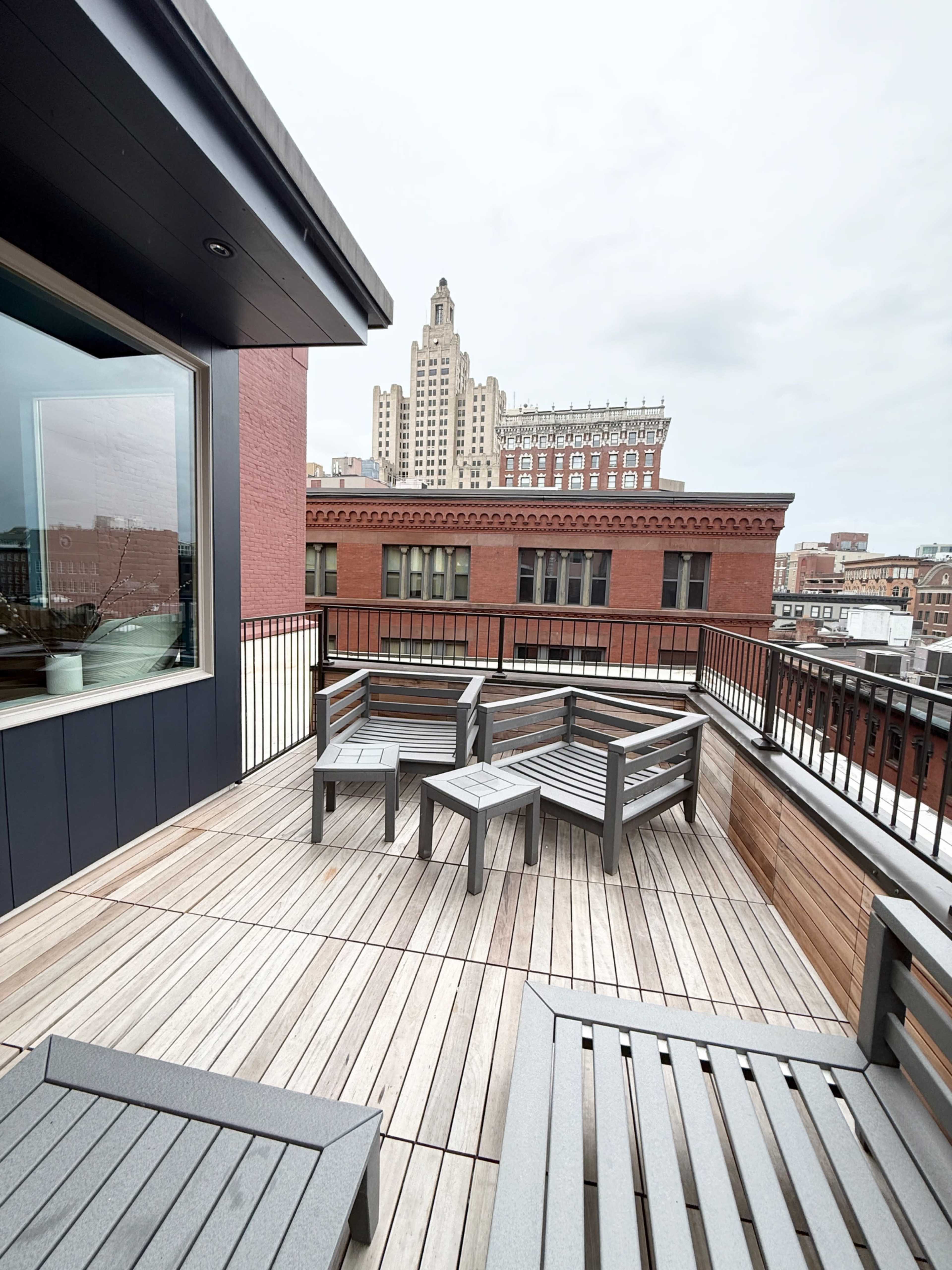 The image shows a rooftop terrace with gray wooden furniture and a view of a tall, historical building in the background.