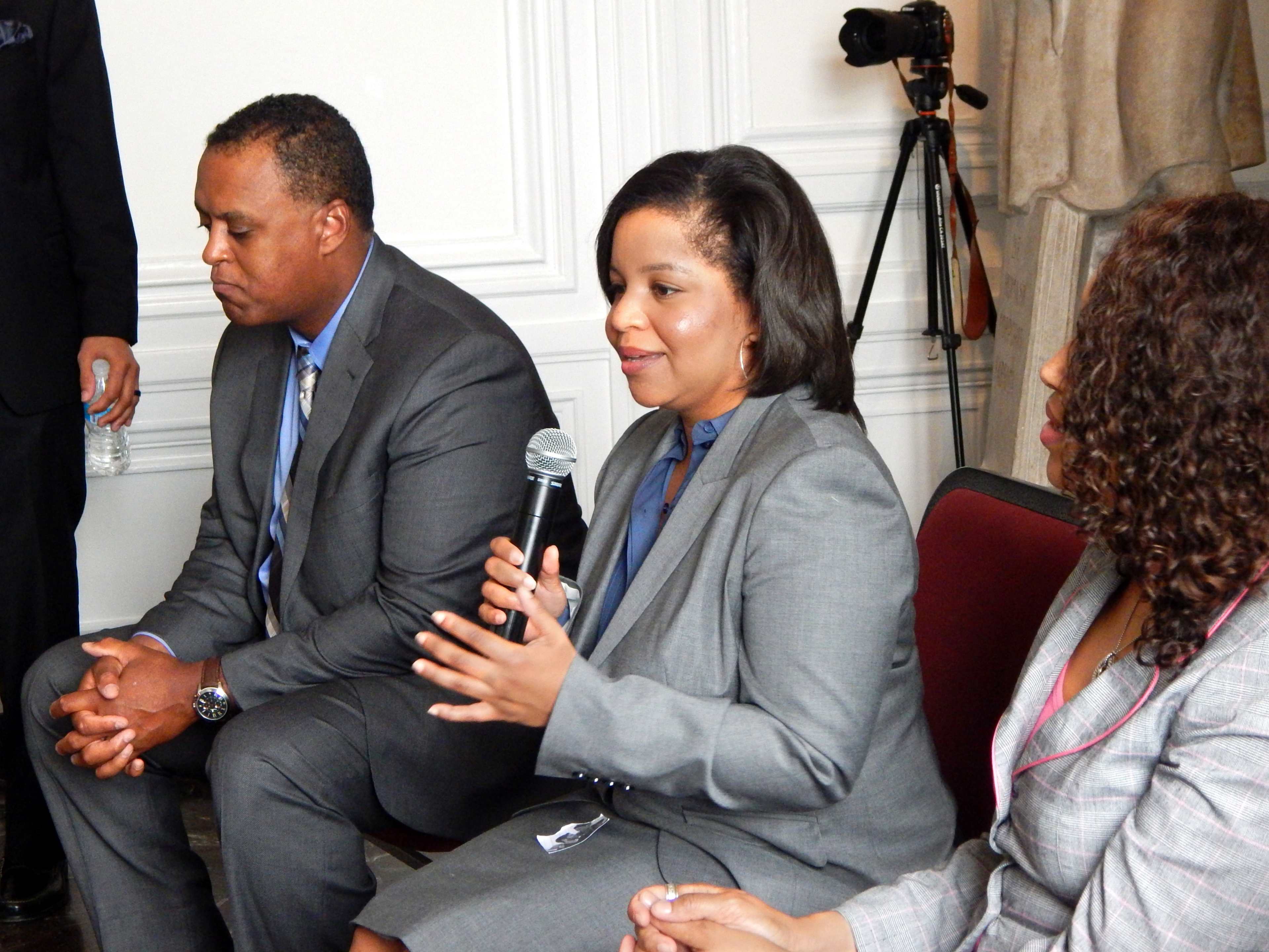 A woman speaks into a microphone while seated between two men in suits at an indoor event.