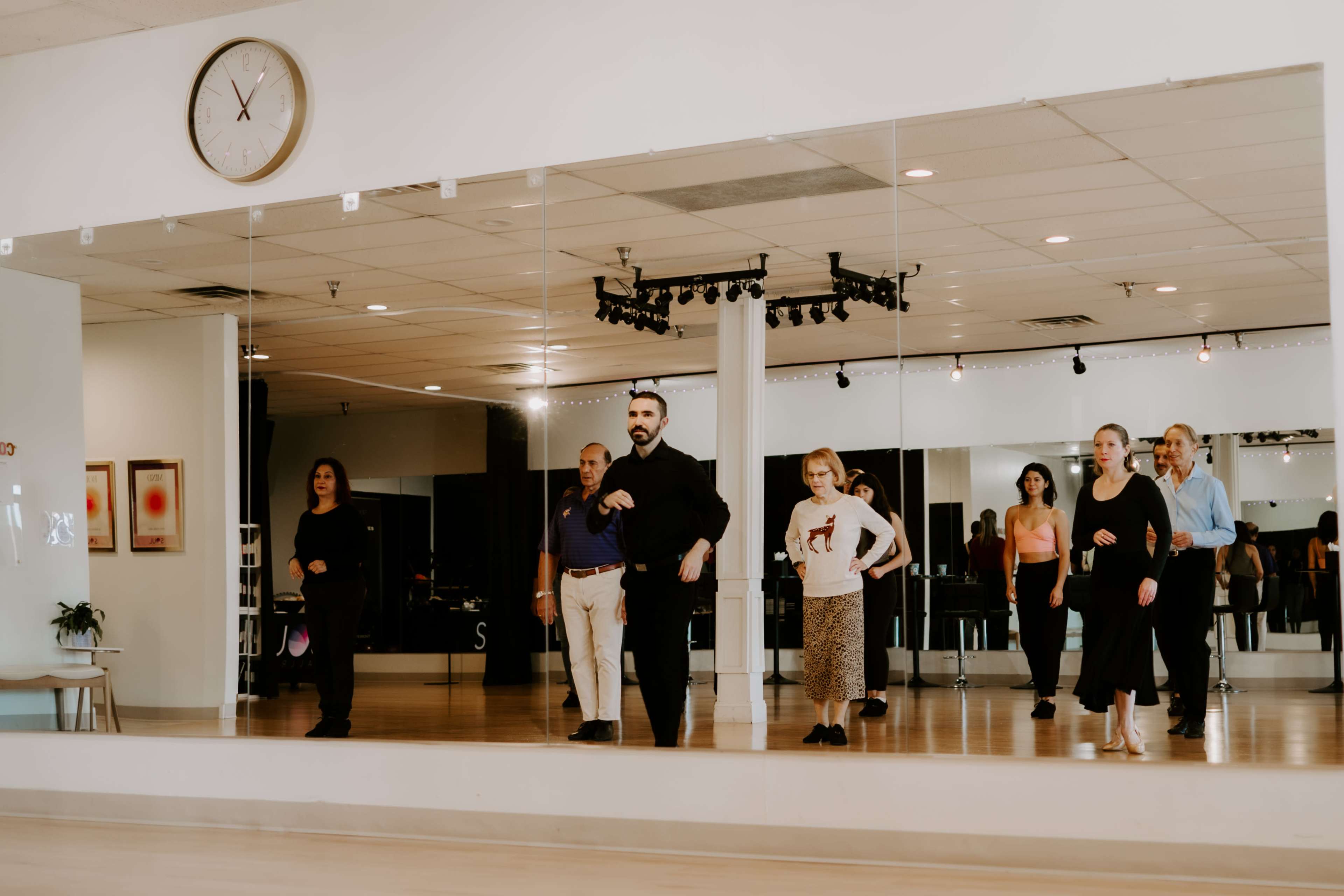 A group of people is practicing dance in a studio with a large mirror and a clock on the wall.
