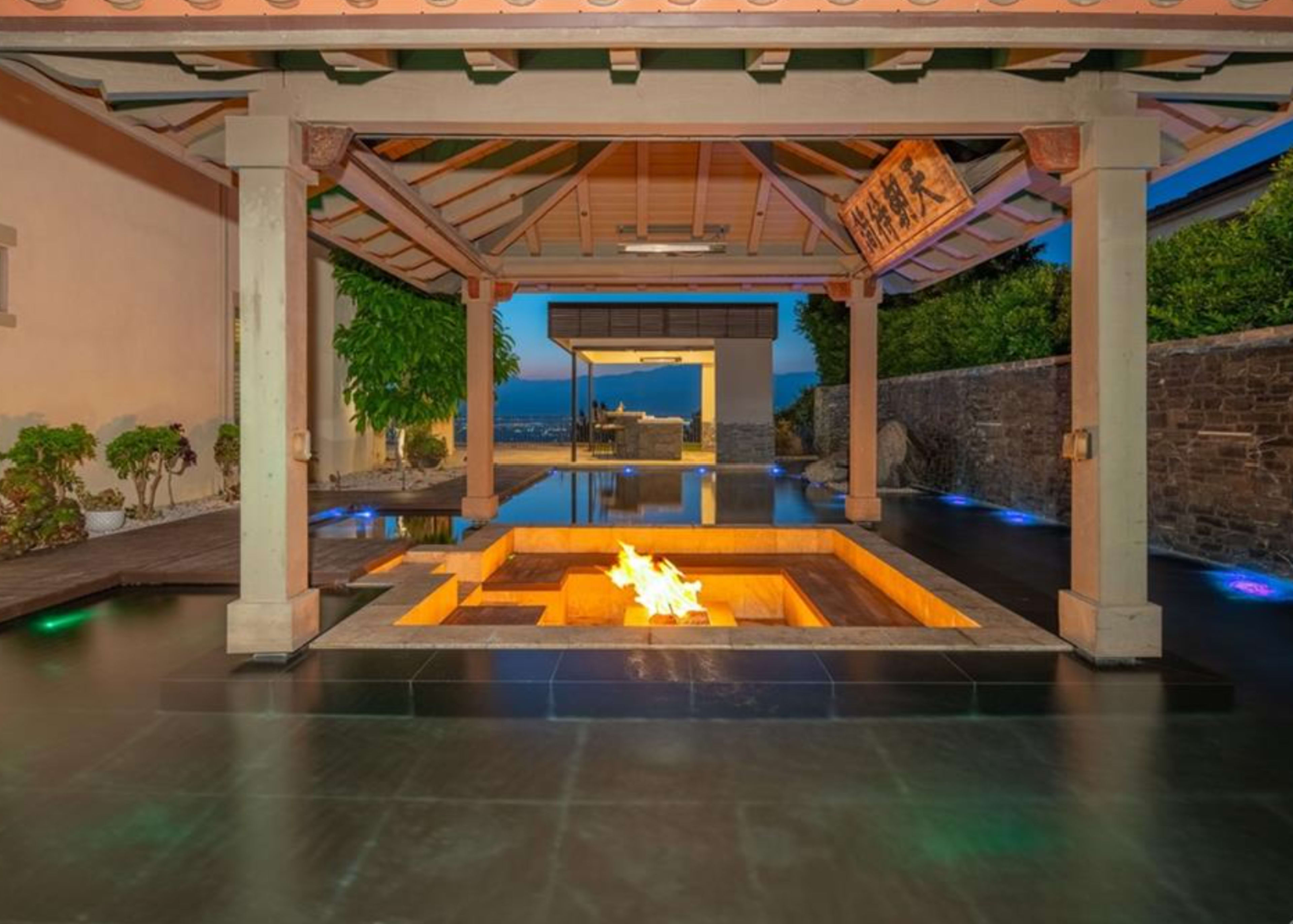 The image shows an outdoor space featuring a pavilion with a square fire pit in the center, surrounded by a reflective pool and illuminated by lights.
