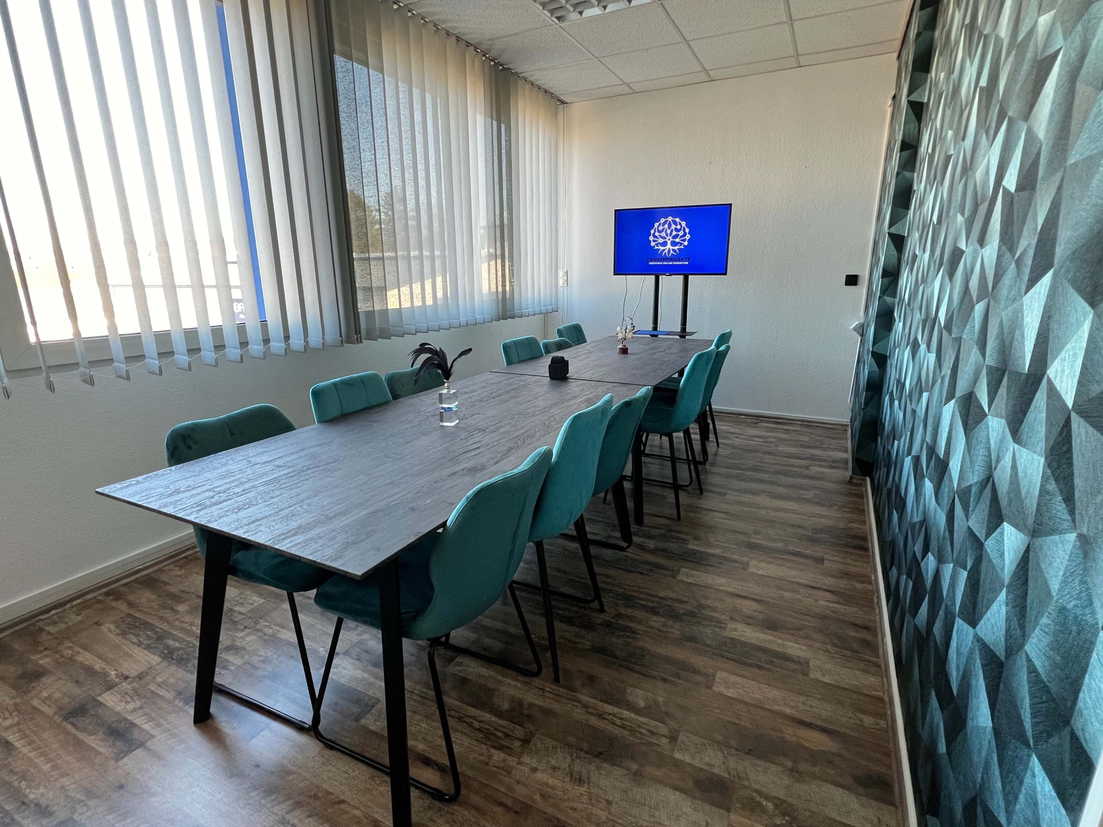 A conference room features a long table surrounded by teal chairs, with a television displaying a logo, and large windows covered by blinds.