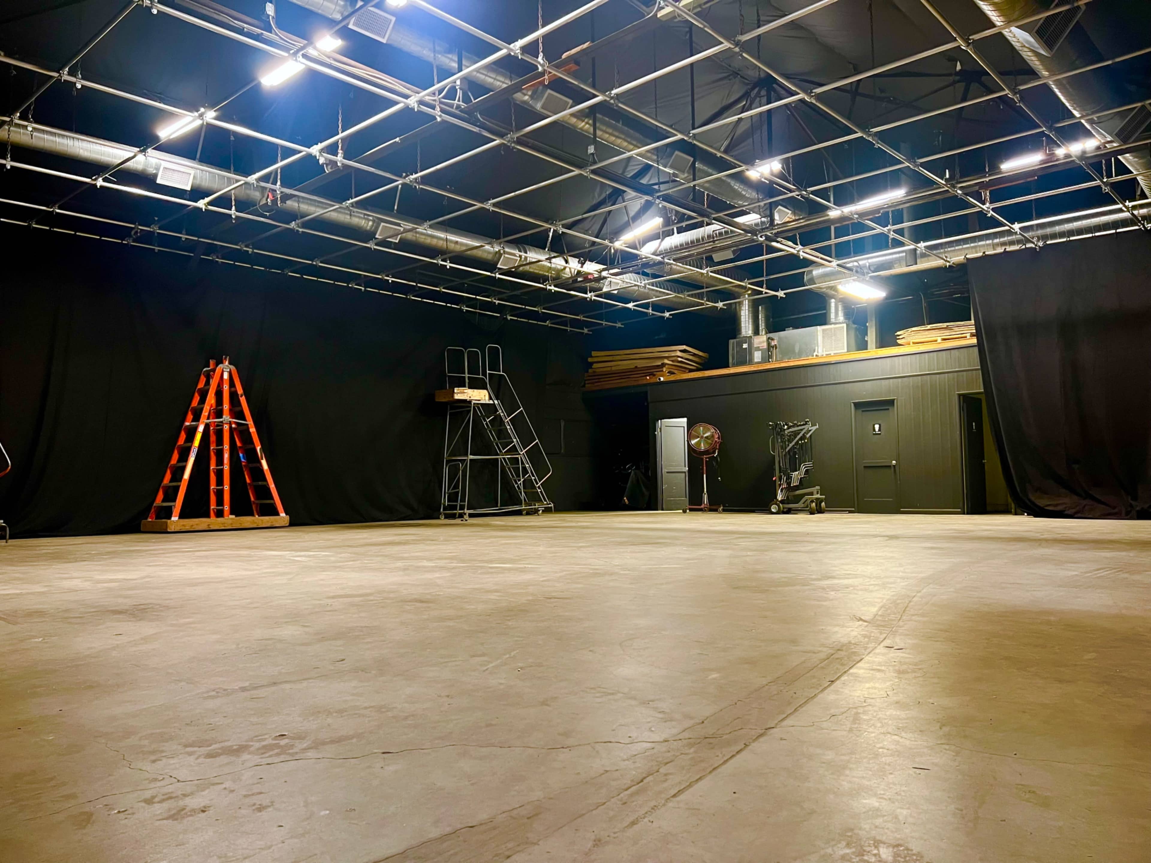 A dimly lit studio space with a bare concrete floor, black curtains lining the walls, and various equipment including a ladder, scaffolding, and a fan.