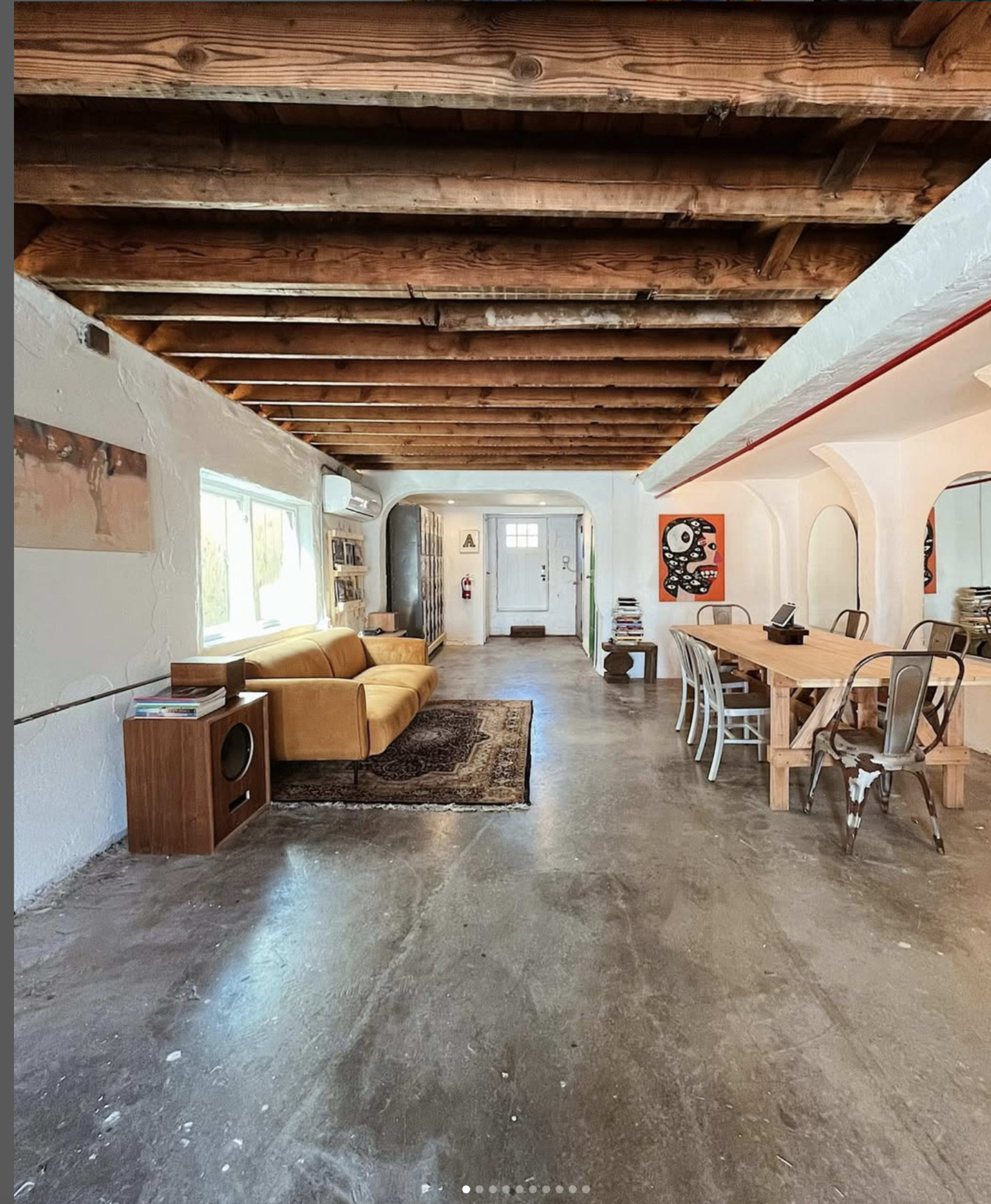 The image shows an open living space with exposed wooden beams, featuring a yellow sofa, a wooden dining table with metal chairs, and a combination of artwork and decor on the walls.