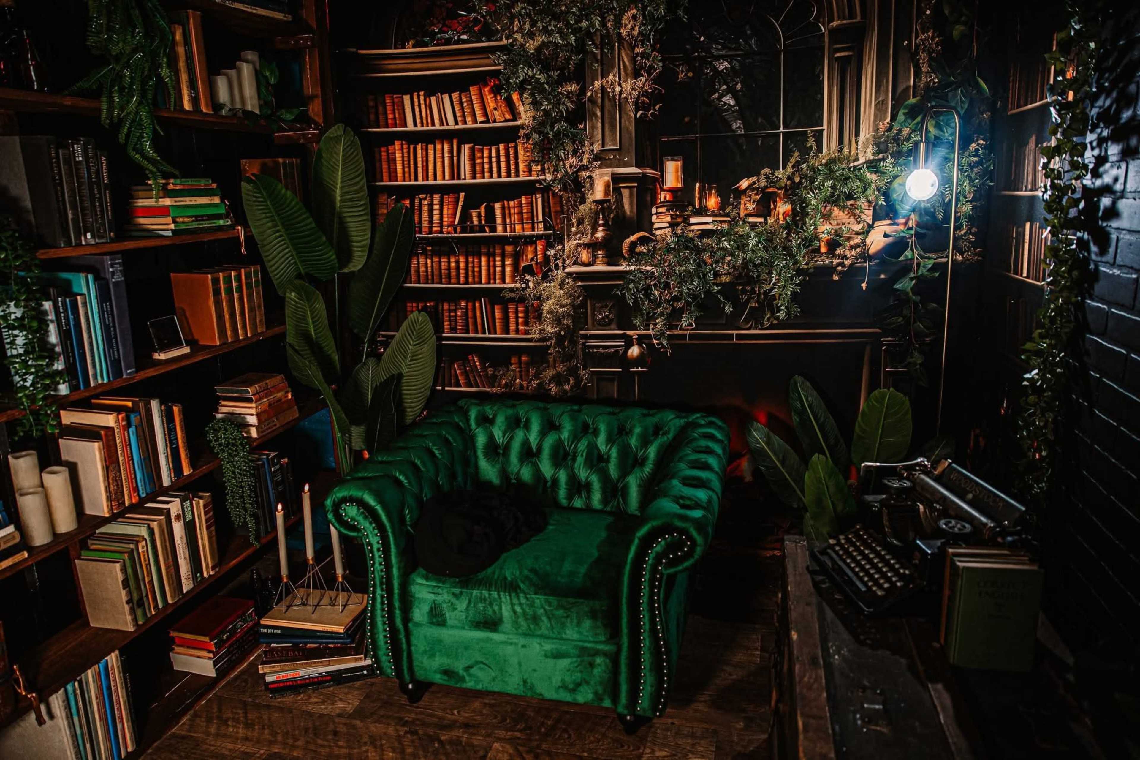 The scene features a green velvet couch in a cozy library setting, surrounded by bookshelves filled with books and decorated with plants.