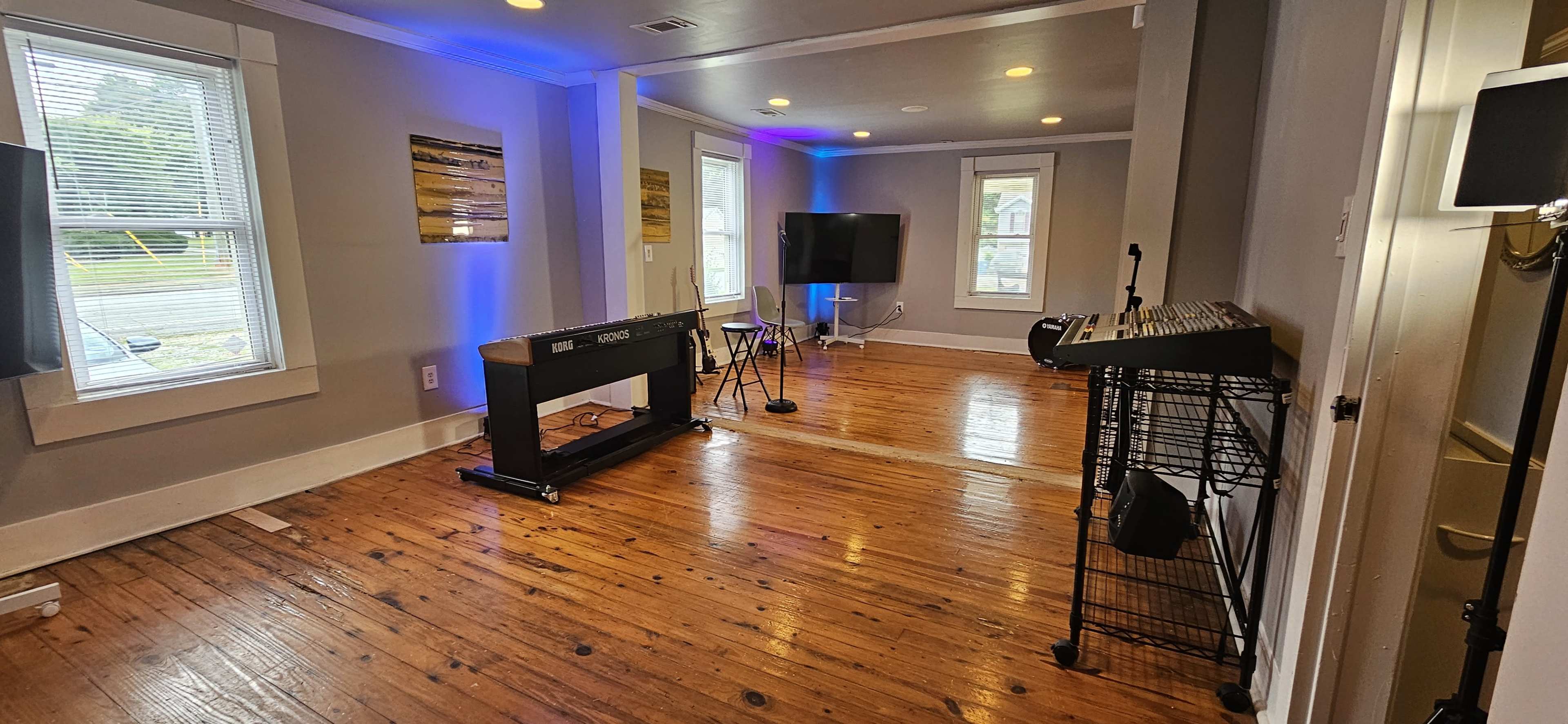 A spacious room with hardwood floors, featuring a keyboard stand, a music stand, and a television setup, illuminated by blue lighting.