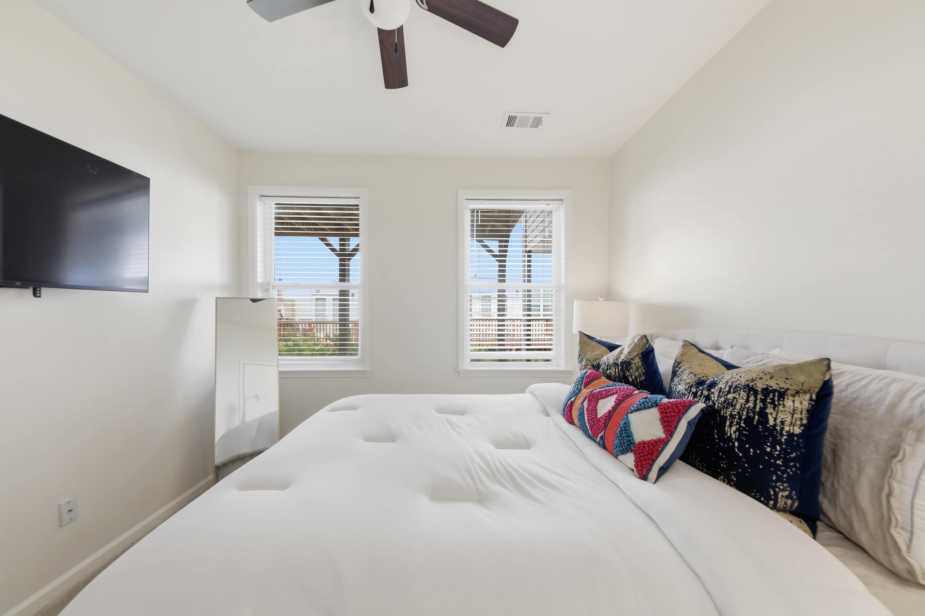 The image shows a well-lit bedroom featuring a bed with decorative pillows, a television mounted on the wall, and two windows with blinds.