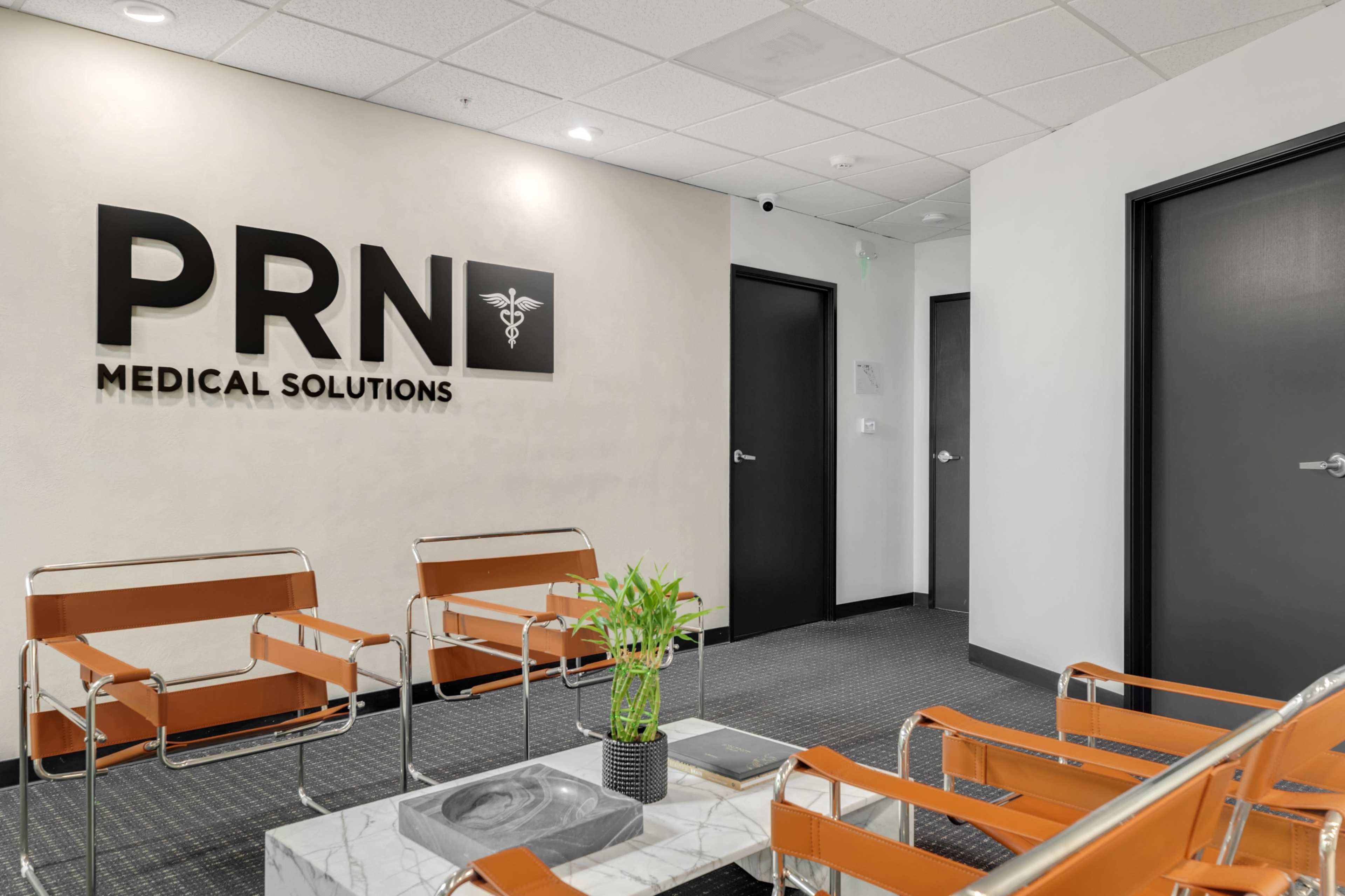 The image shows a modern waiting area with orange chairs, a marble coffee table, and a sign that reads "PRN Medical Solutions" on the wall.