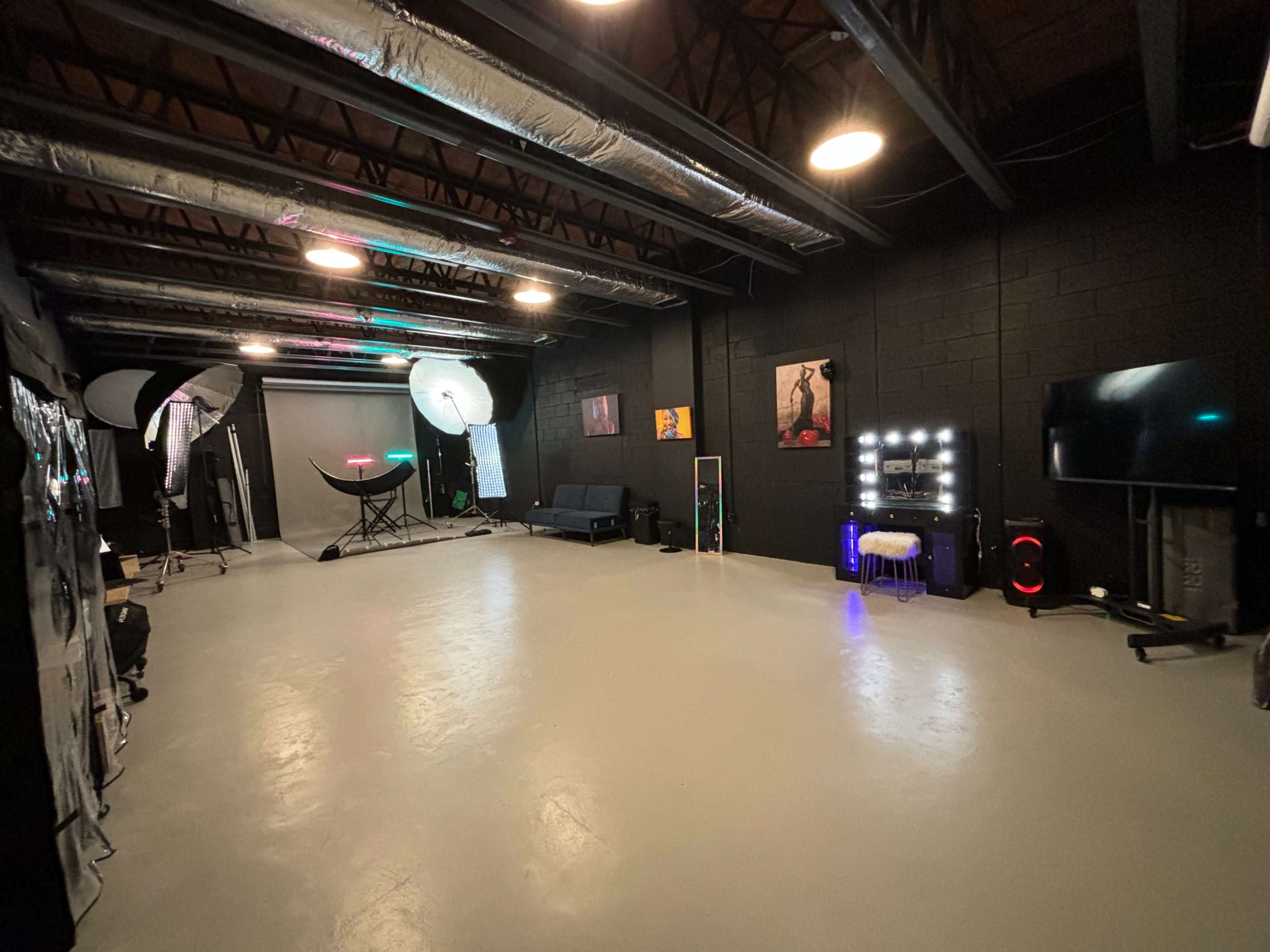 The image shows a spacious studio with lighting equipment, a couch, and a large screen along one wall, set against black-painted walls and a polished floor.