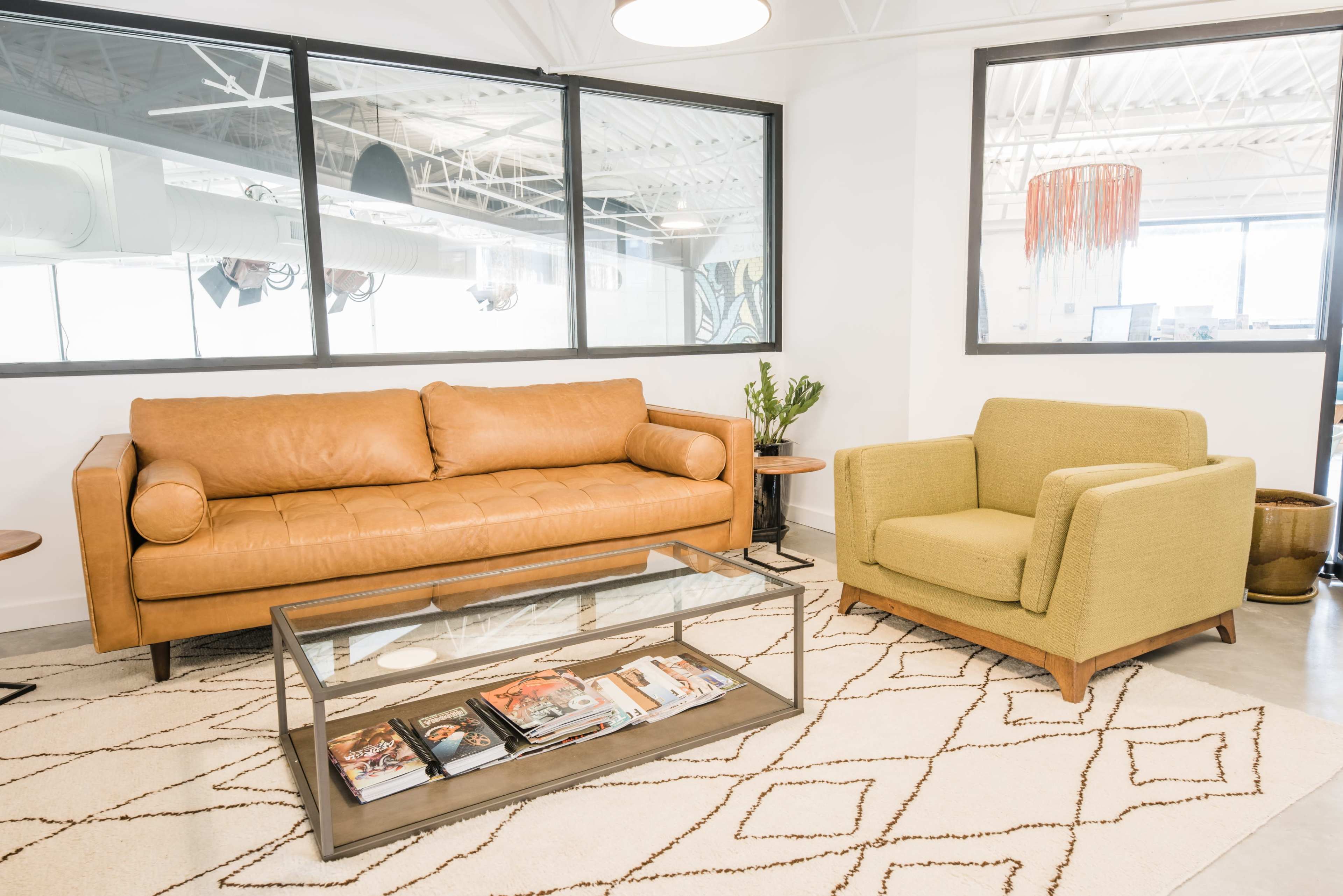 A modern lounge area with a brown leather sofa, a green armchair, a glass coffee table, and large windows allowing natural light.