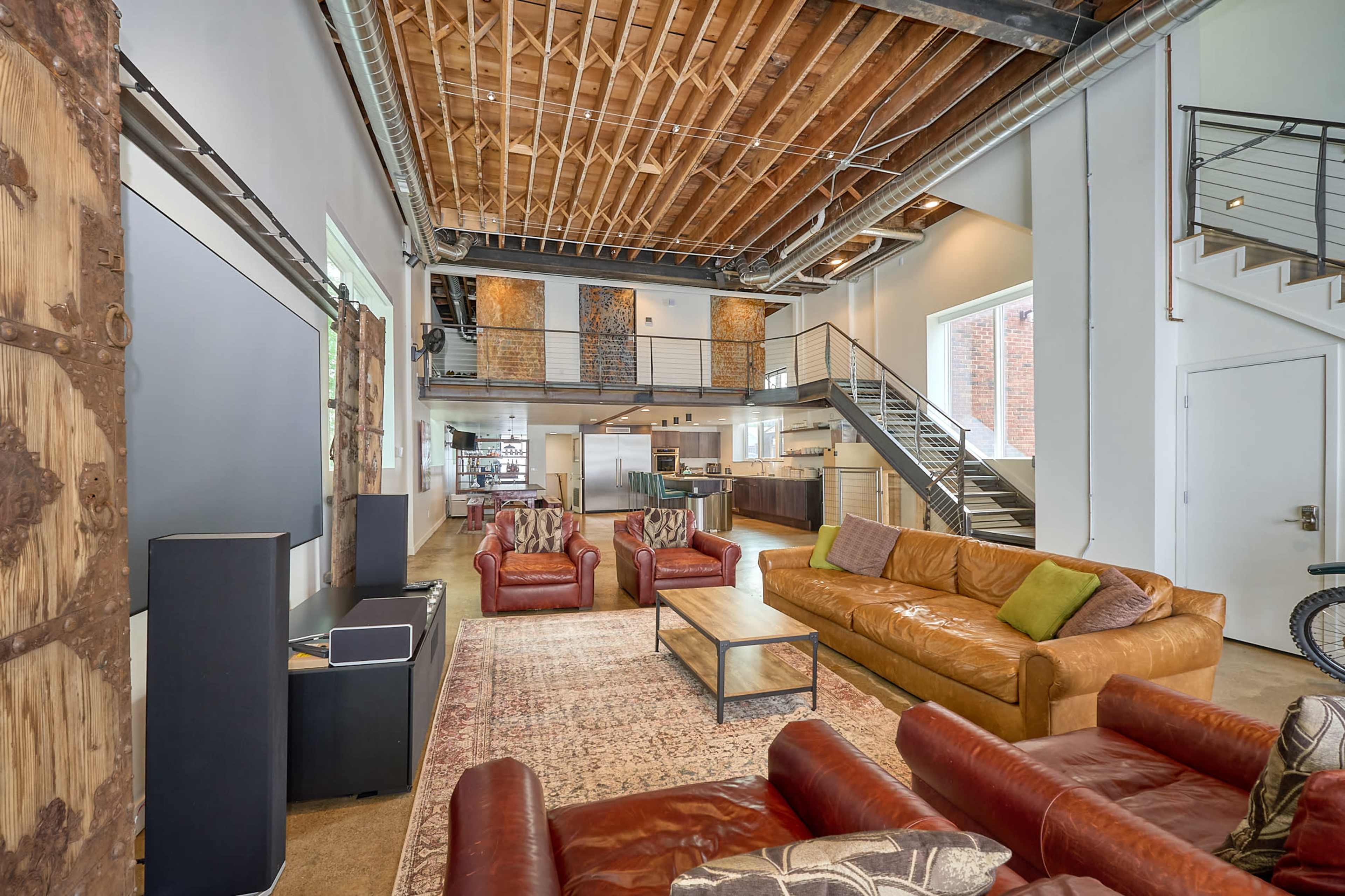 A spacious, modern living area with exposed wooden beams, multiple seating options, and a visible kitchen area in a loft-style design.