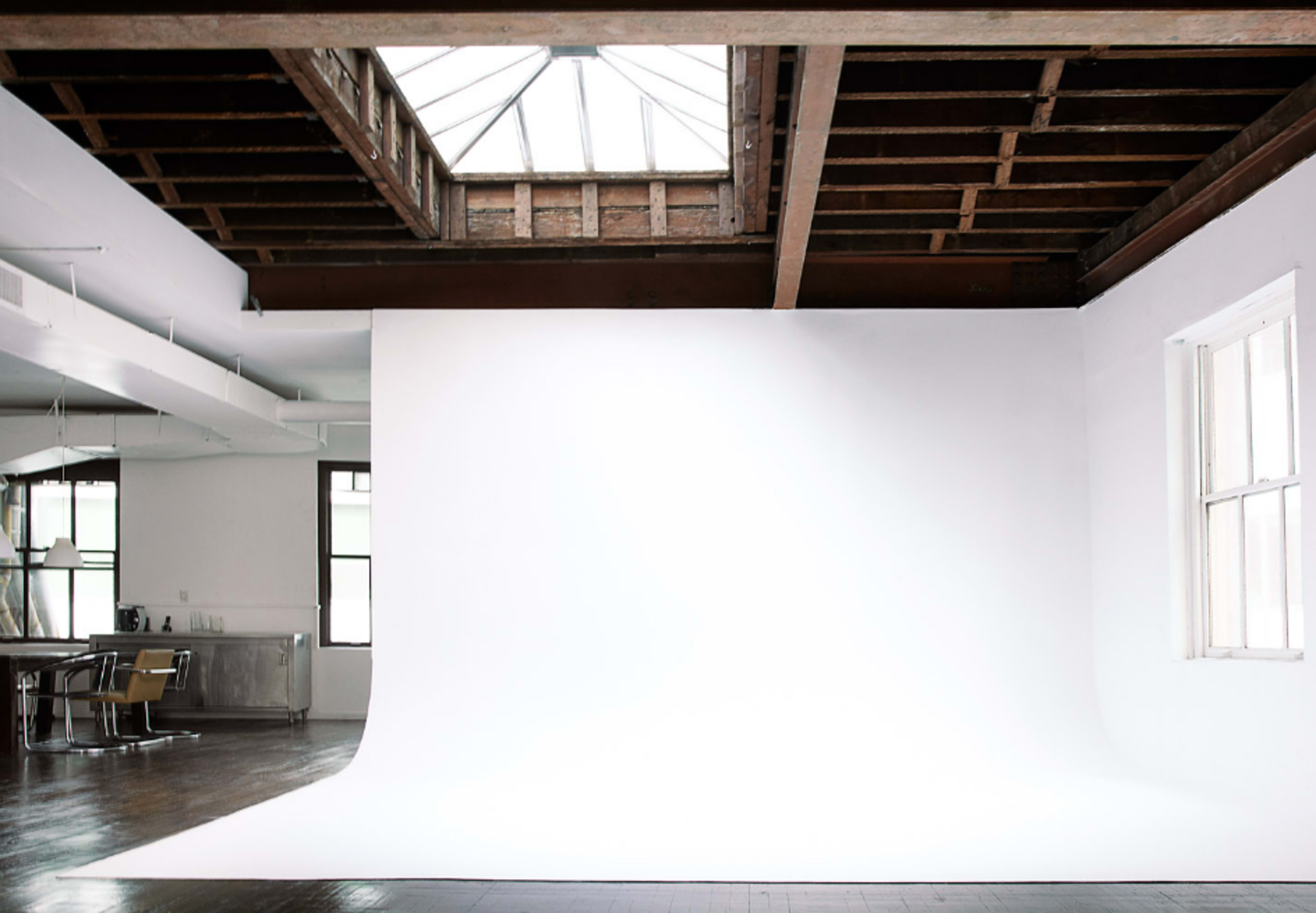A spacious, minimalist studio with a large white backdrop and exposed wooden beams, featuring a skylight above.