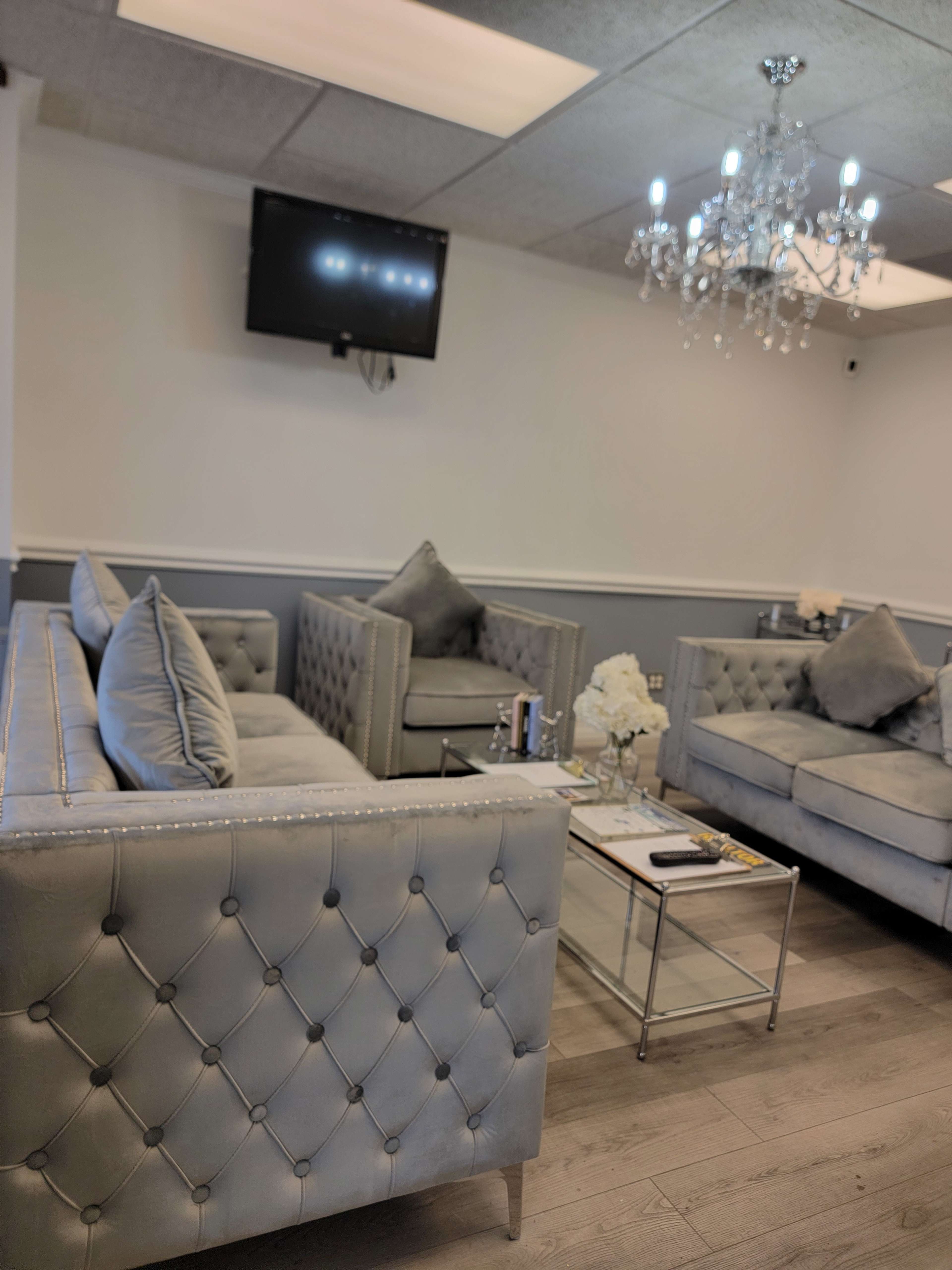 The image shows a modern lounge area with tufted gray sofas, a glass coffee table, and a chandelier, accompanied by a wall-mounted television.