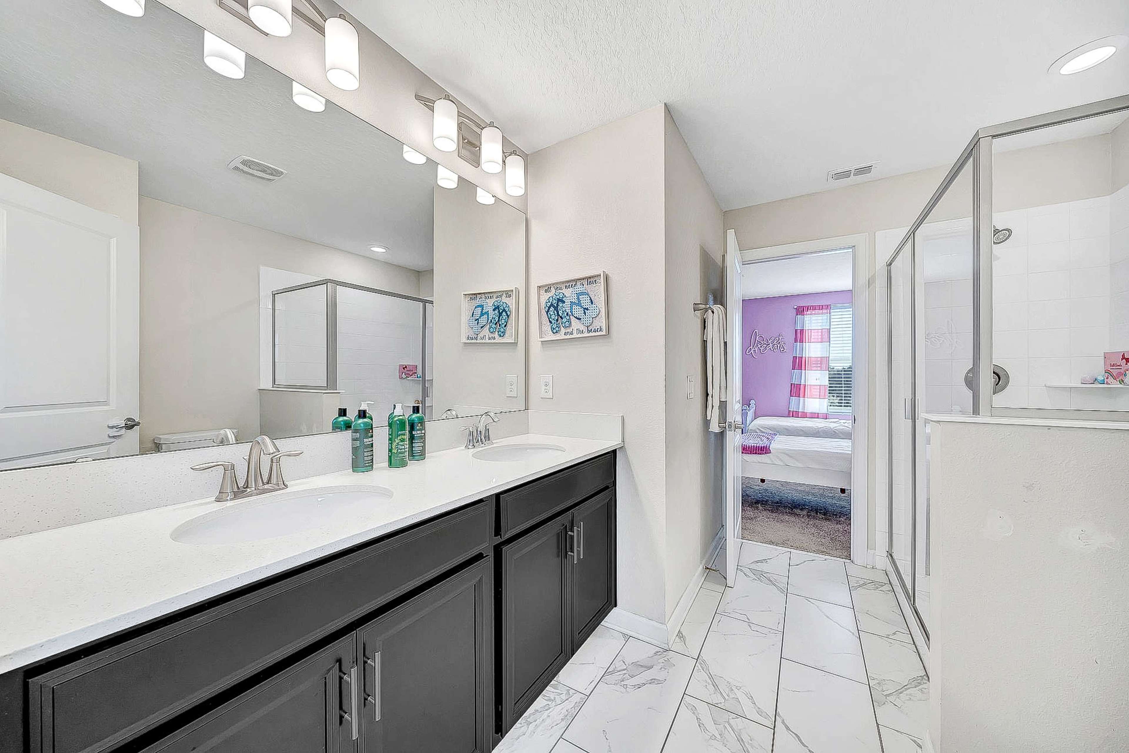 A modern bathroom with dual sinks, a large mirror, and a glass shower, featuring light-colored walls and marble flooring.