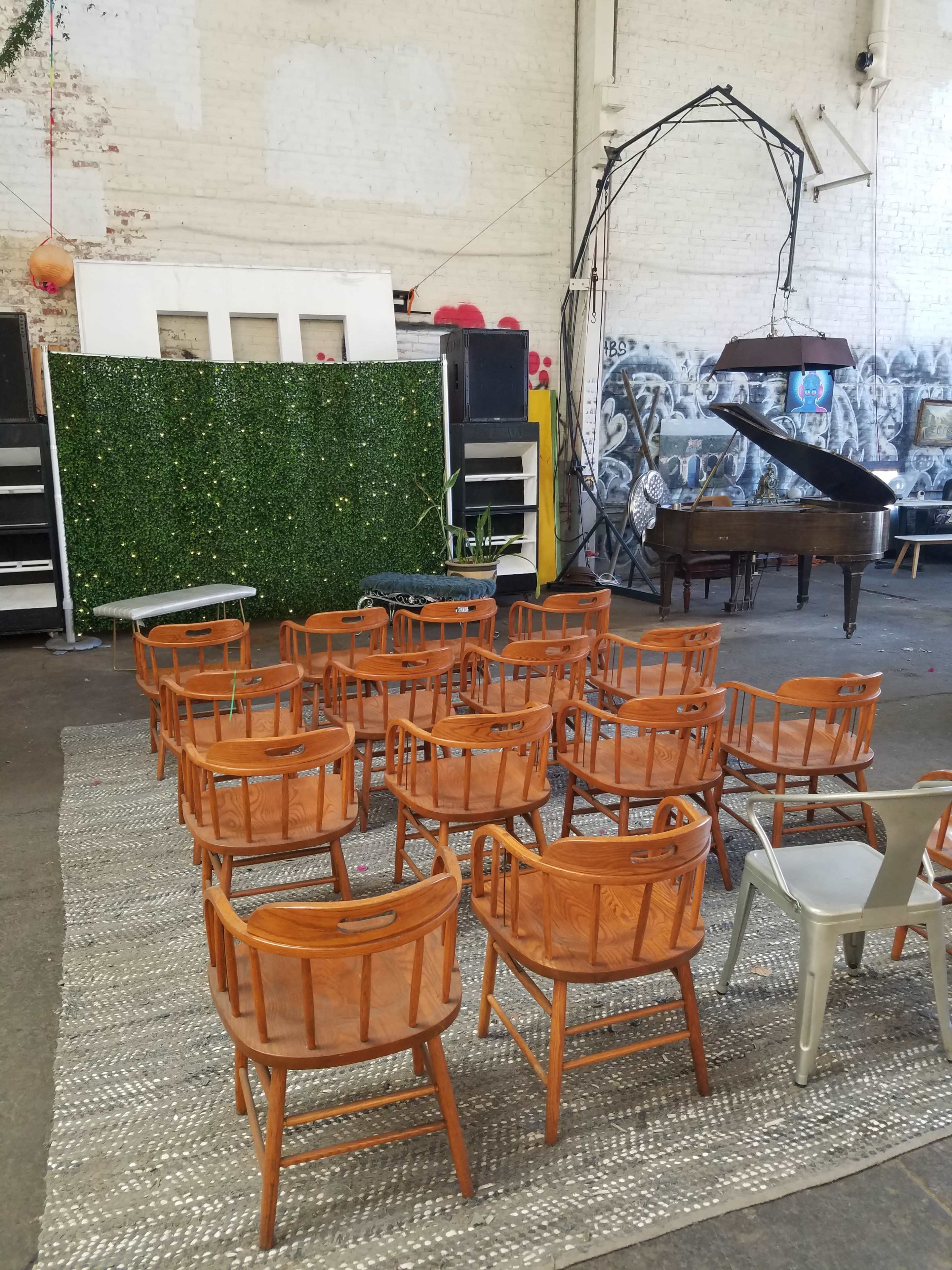 The image shows an arrangement of wooden chairs facing a green wall decoration, with a grand piano and sound equipment in the background of a spacious indoor venue.