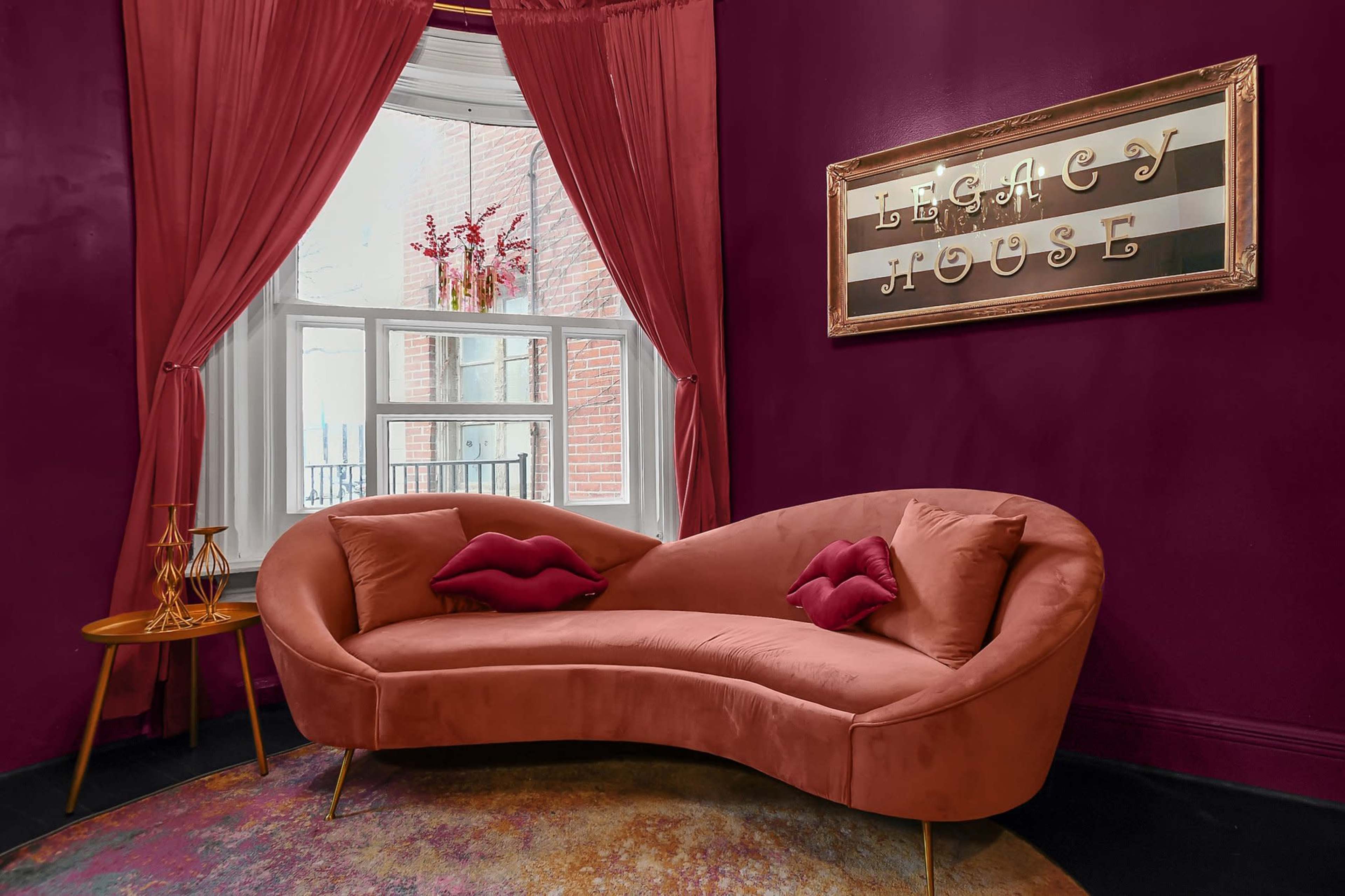 A curved, pink velvet sofa with decorative pillows, positioned next to a window adorned with red curtains in a room featuring a deep purple wall and a framed sign that reads "LEGACY HOUSE."