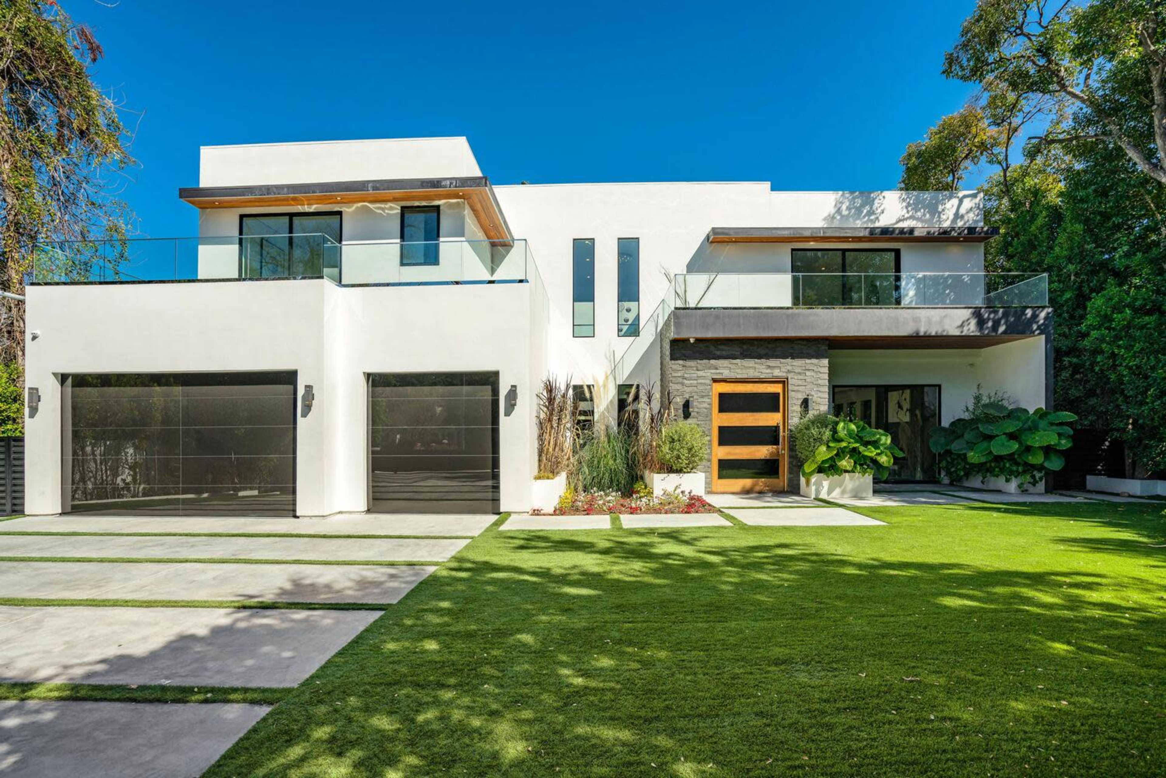 A modern two-story house features large windows, a combination of white and stone exterior, and a manicured lawn with a symmetrical driveway.