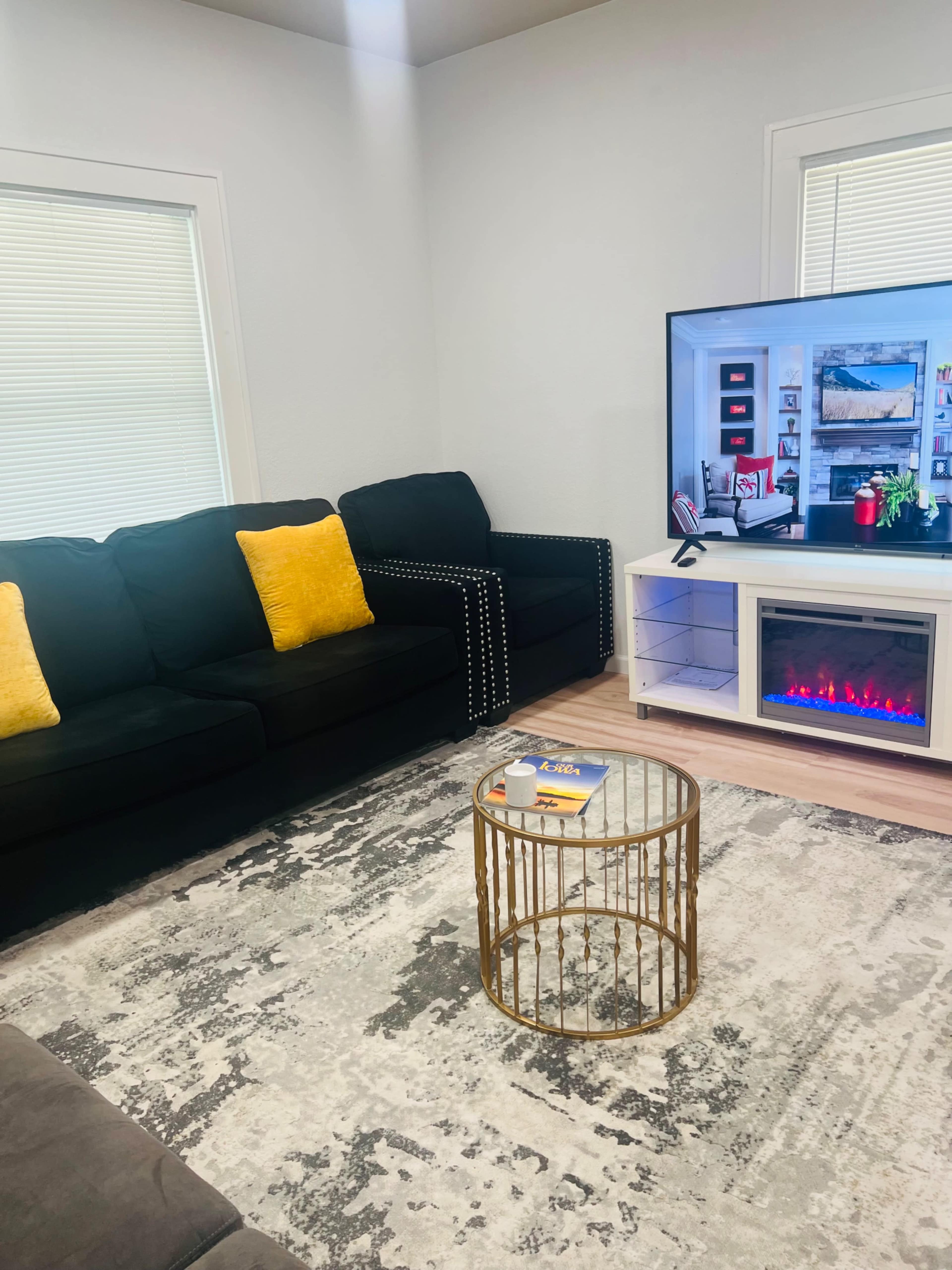 A living room features a black couch and armchairs, a patterned area rug, a glass coffee table, and a television mounted on the wall.