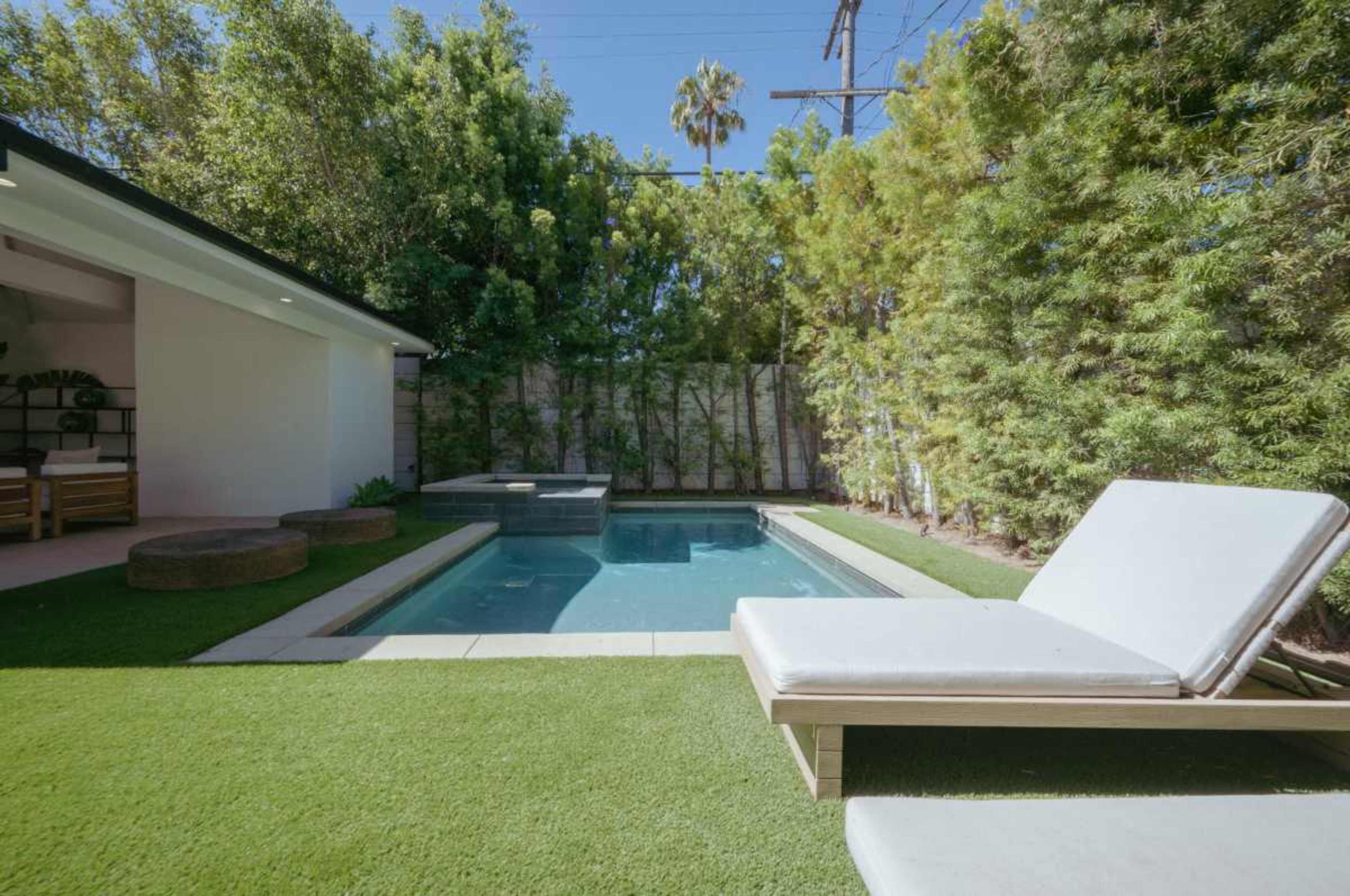 A modern swimming pool is surrounded by lush greenery and features a built-in hot tub, with a lounge chair positioned nearby.