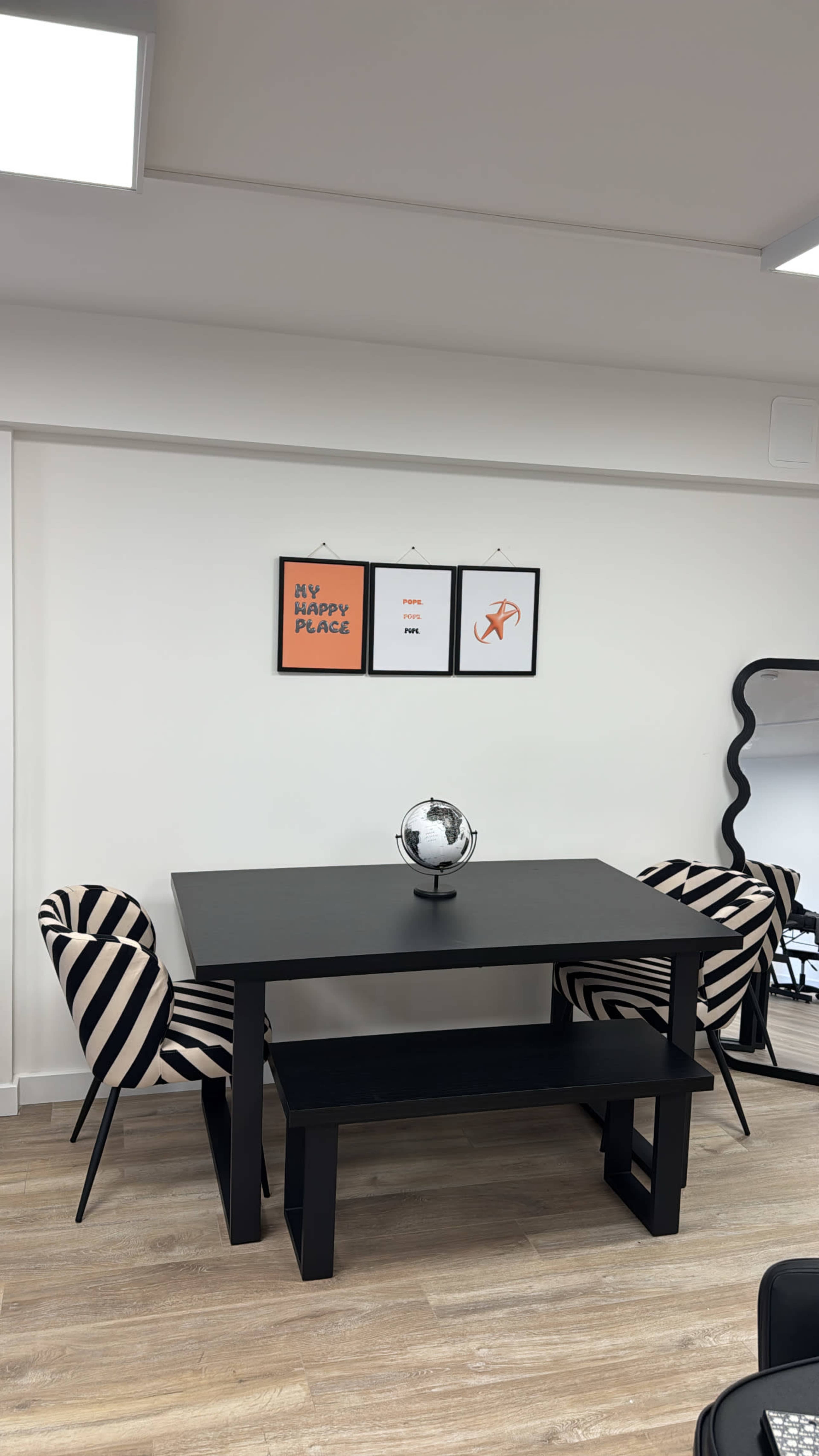 The image depicts a modern dining area featuring a black table with two striped chairs, a small globe on the table, and framed artwork on the wall.