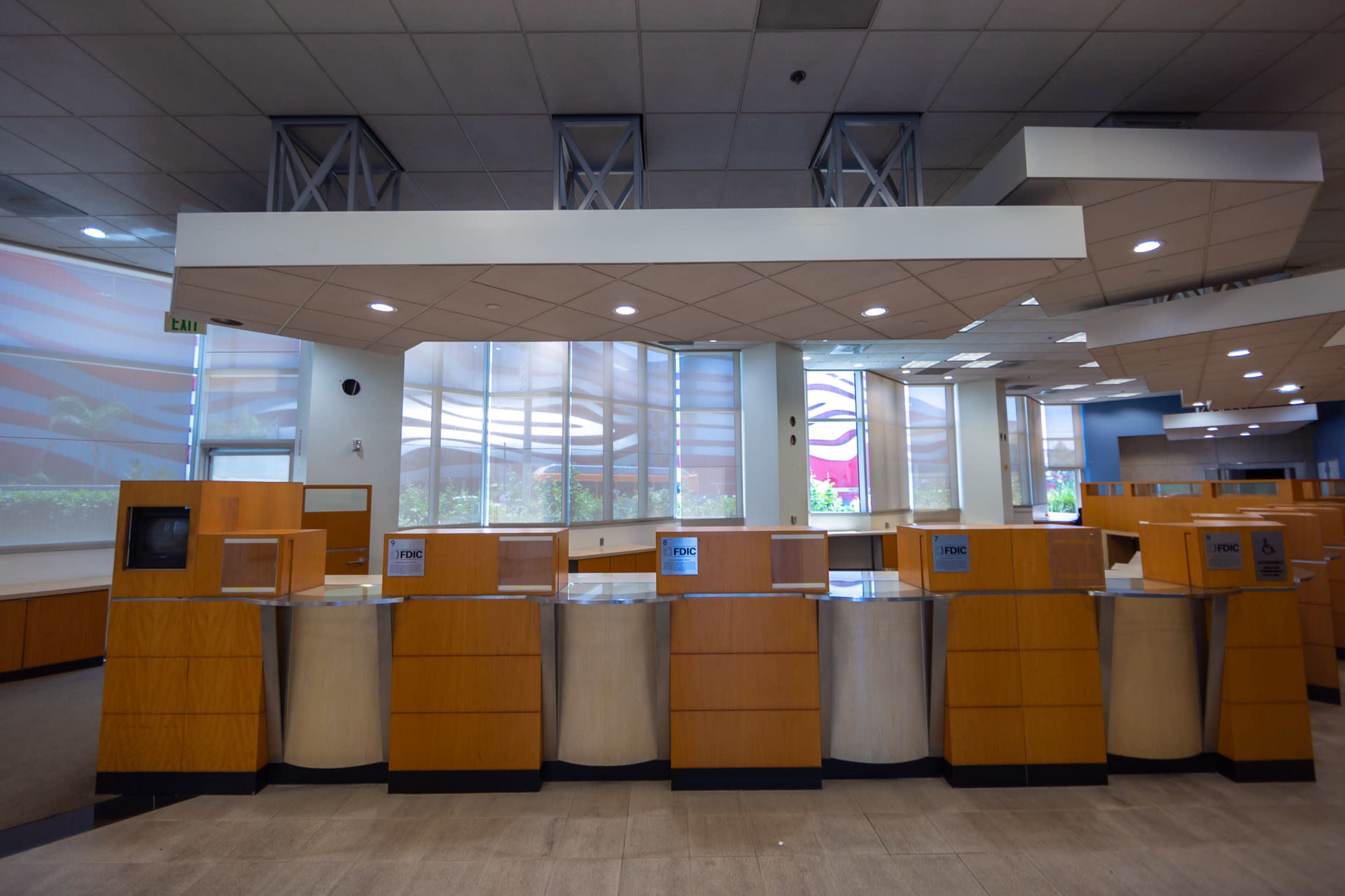 The image shows a spacious, modern lobby with several reception desks arranged in a row, featuring wooden finishes and large windows revealing a view of the exterior.