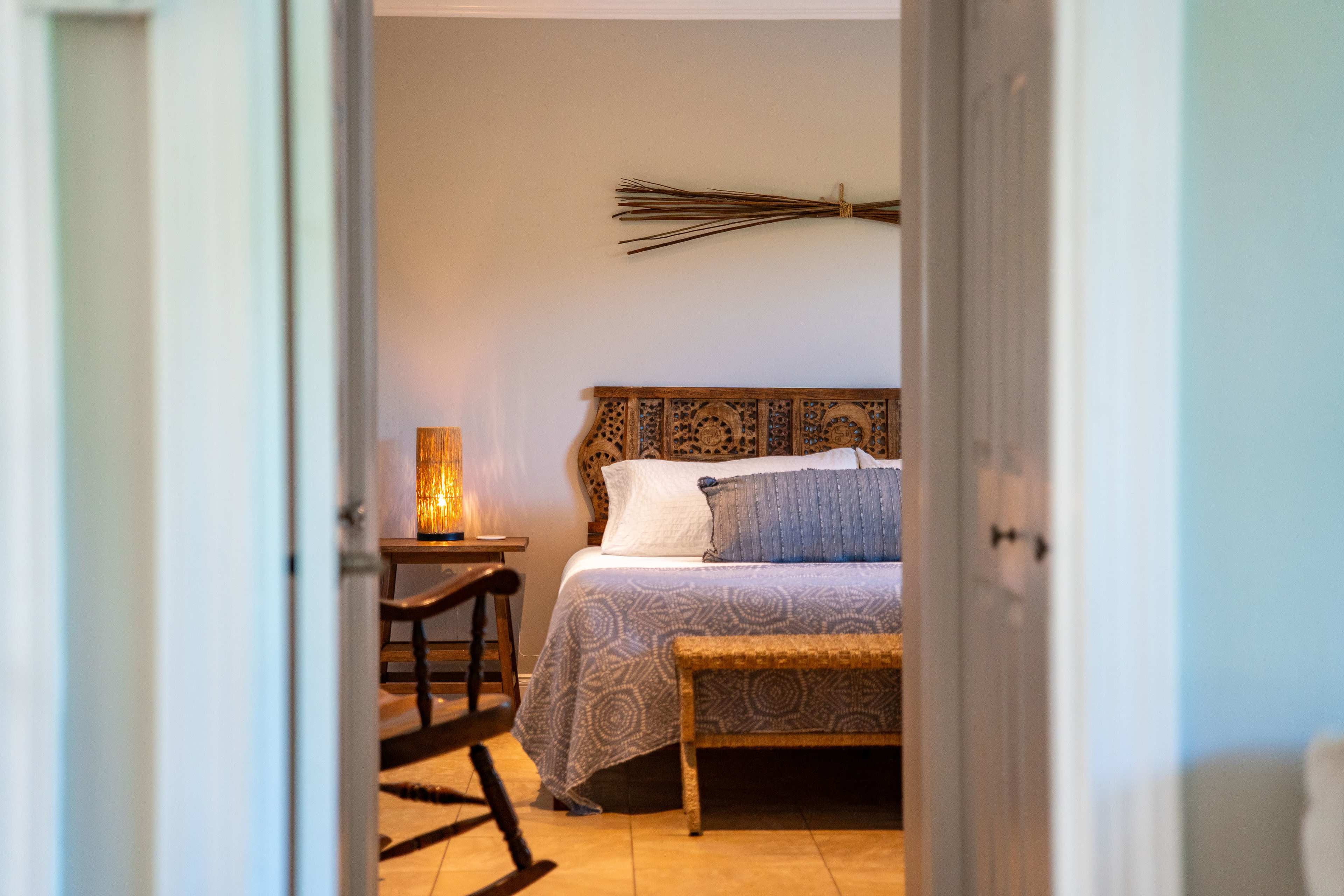 A neatly arranged bedroom features a wooden bed with a decorative headboard, a woven bench at the foot, and a lamp casting warm light.