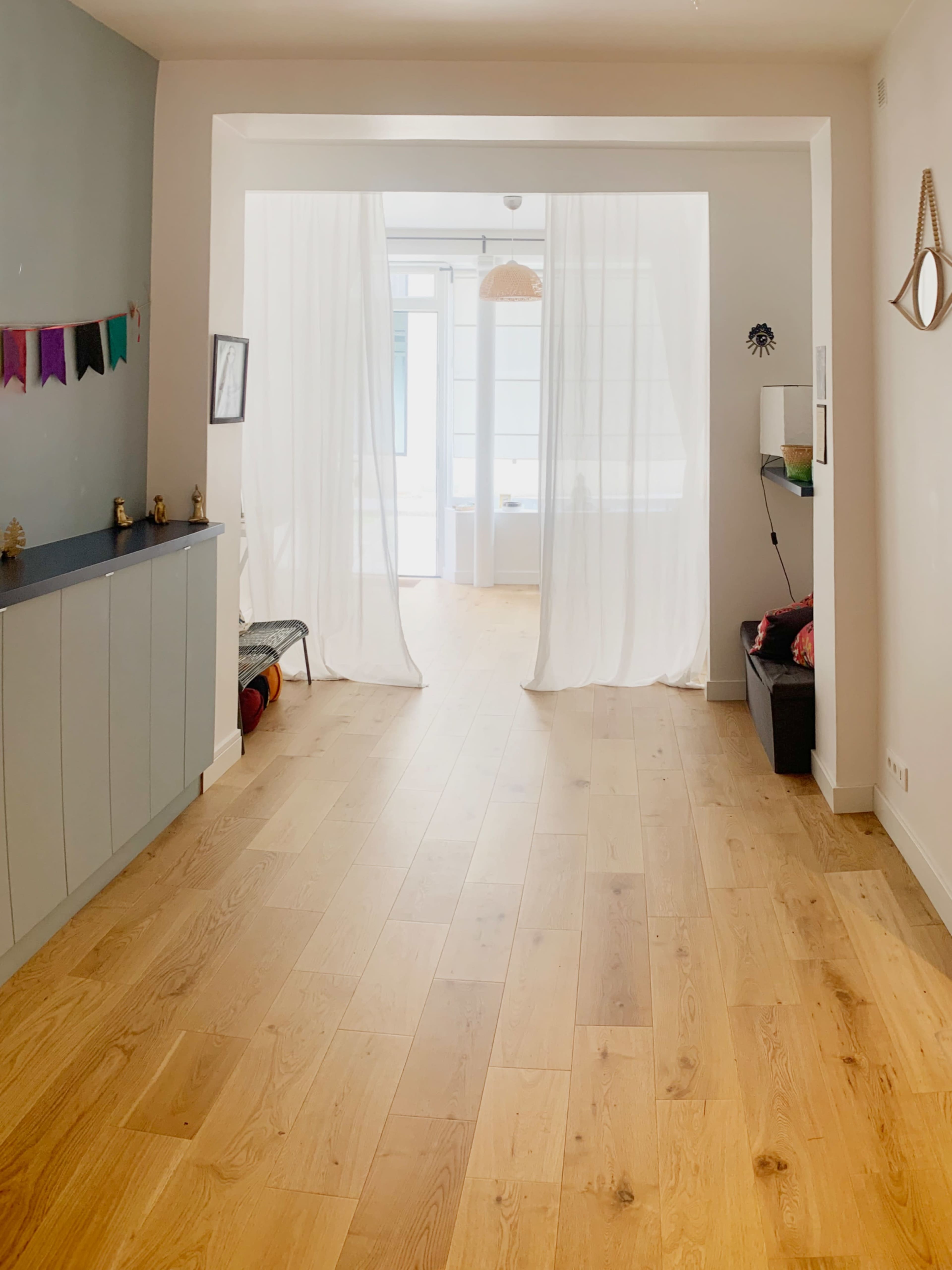 The image shows a bright interior hallway with wooden flooring, light curtains dividing the space, and minimal furniture against the walls.