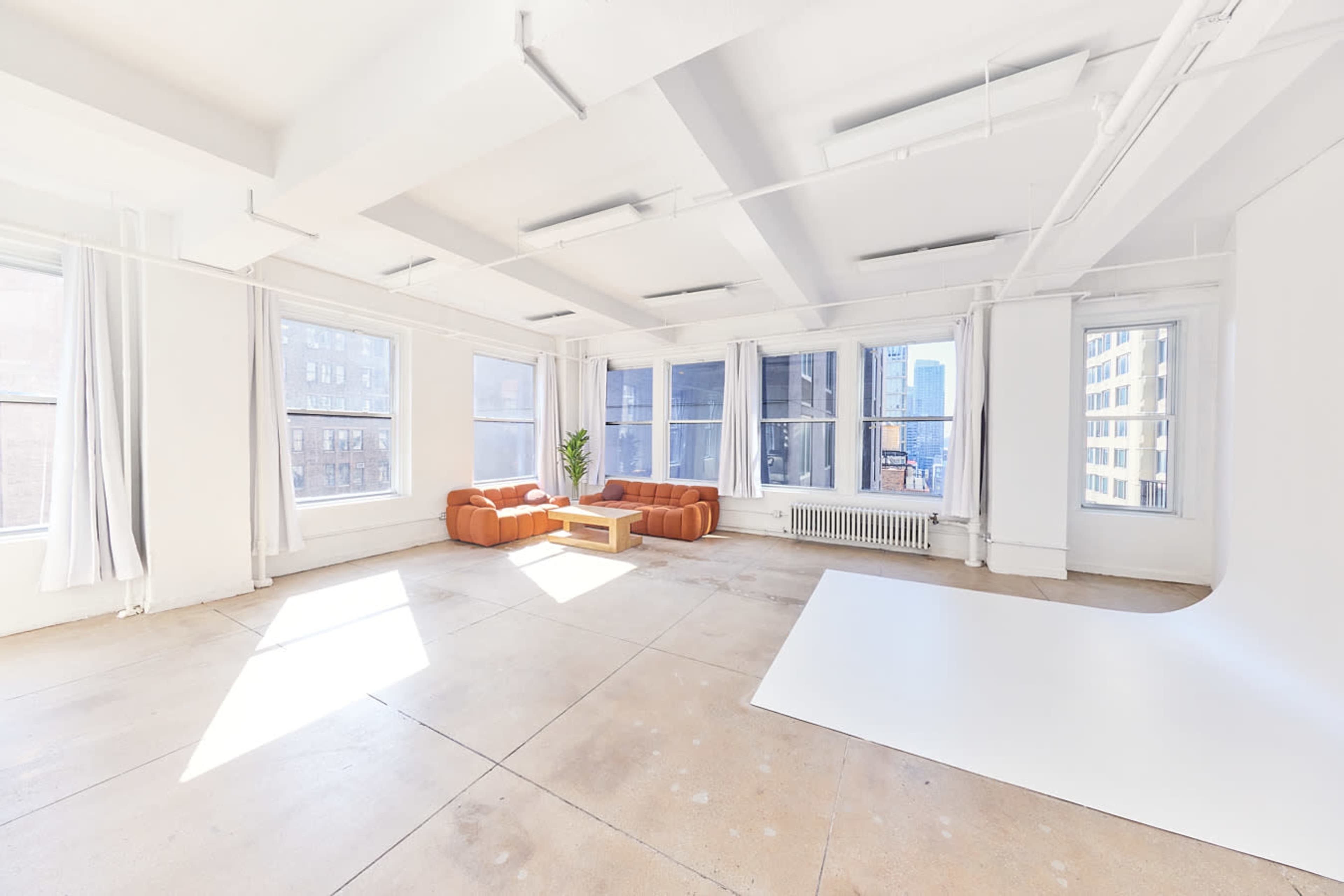 A spacious, well-lit studio features large windows, a pair of brown sofas, a wooden coffee table, and a minimalist white backdrop.