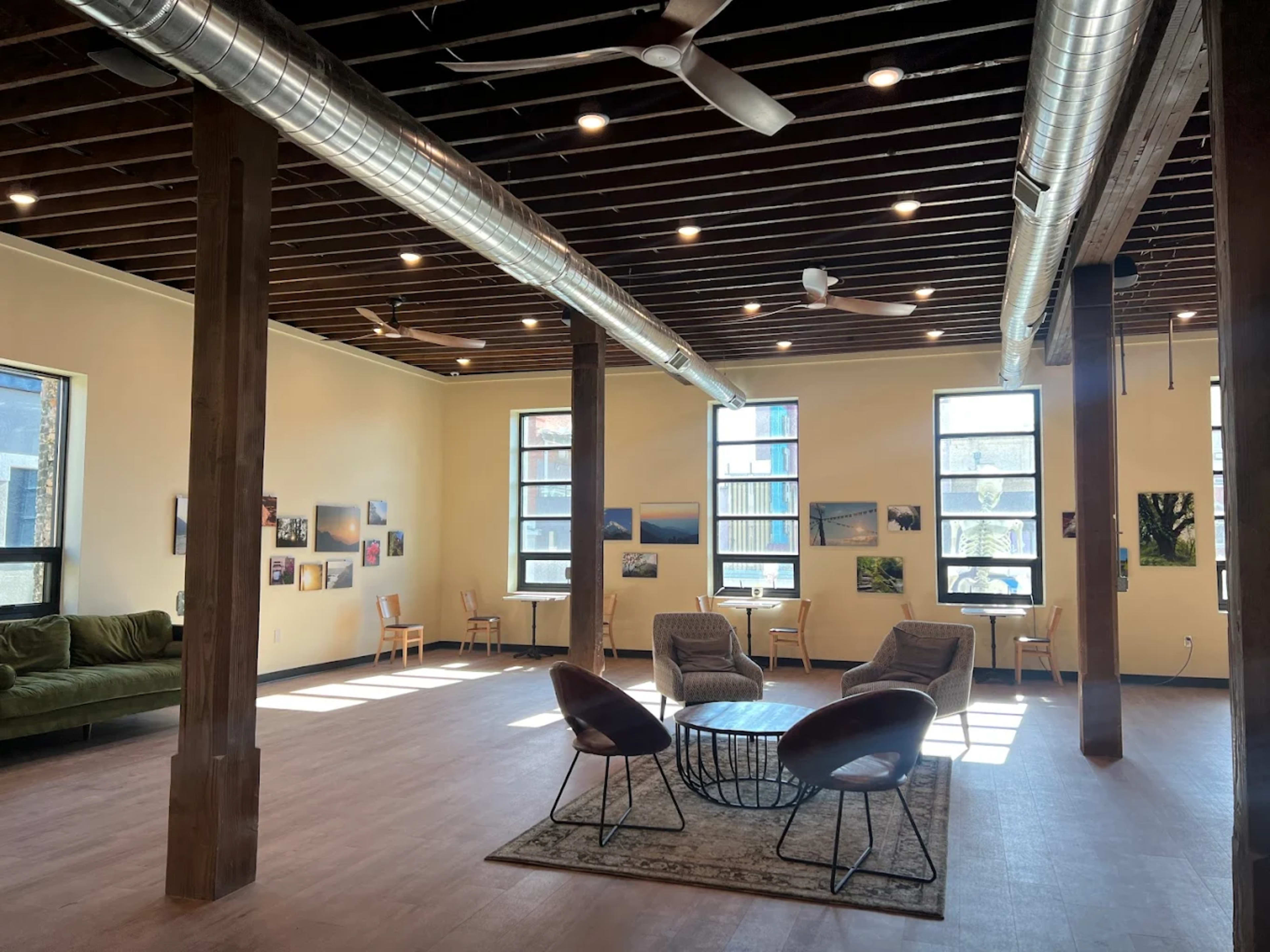 The image shows a spacious, well-lit interior with high ceilings, wooden beams, and a mixture of seating arrangements including a green sofa and several chairs around a coffee table.
