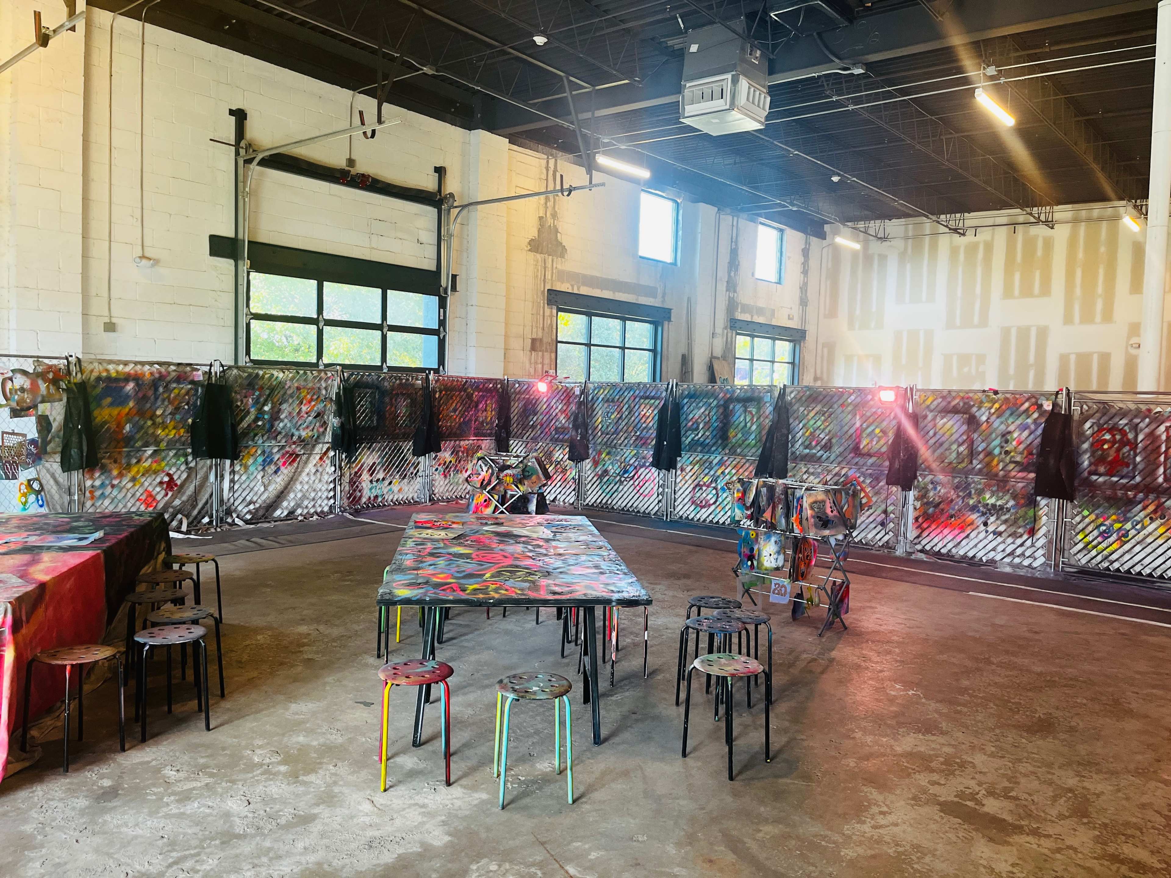 An industrial-style room with a concrete floor, several brightly painted chairs, a table covered in colorful designs, and walls adorned with graffiti and art installations.