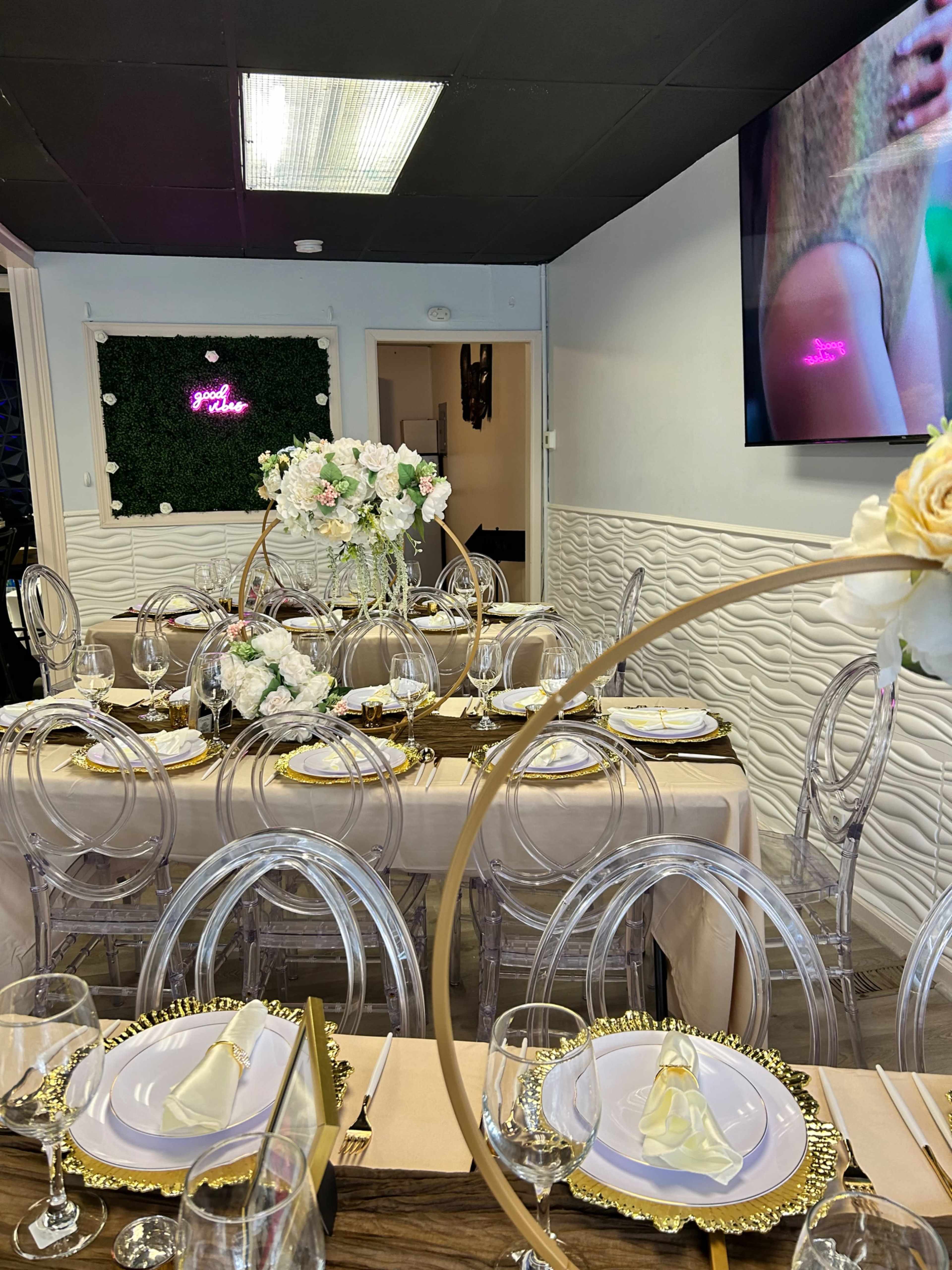 A dining area set with clear chairs, elegant table settings, and floral centerpieces, with a decorative wall featuring greenery and a neon sign.