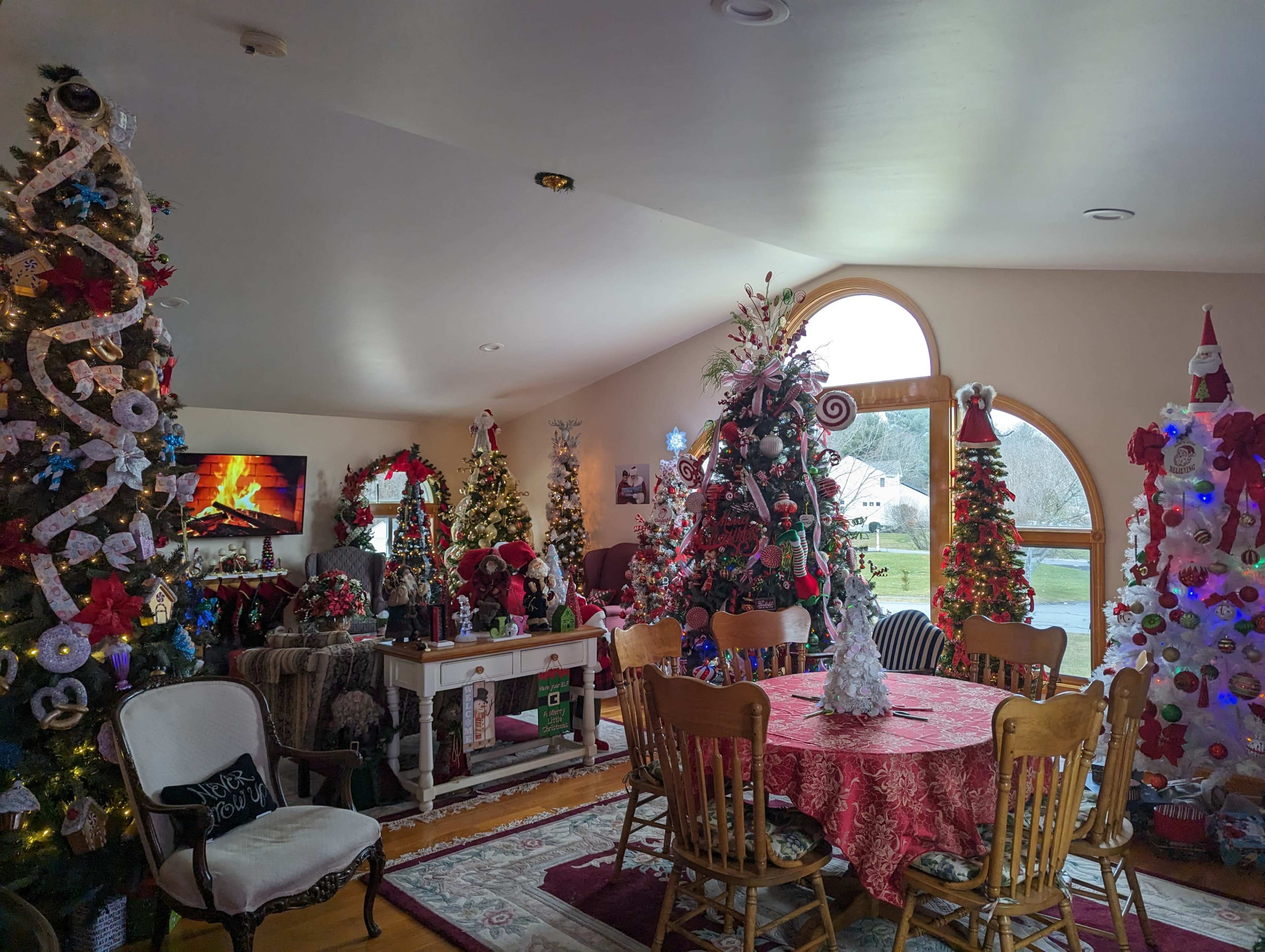 A festive room decorated with several Christmas trees, a dining table covered with a red tablecloth, and visible holiday decorations throughout the space.