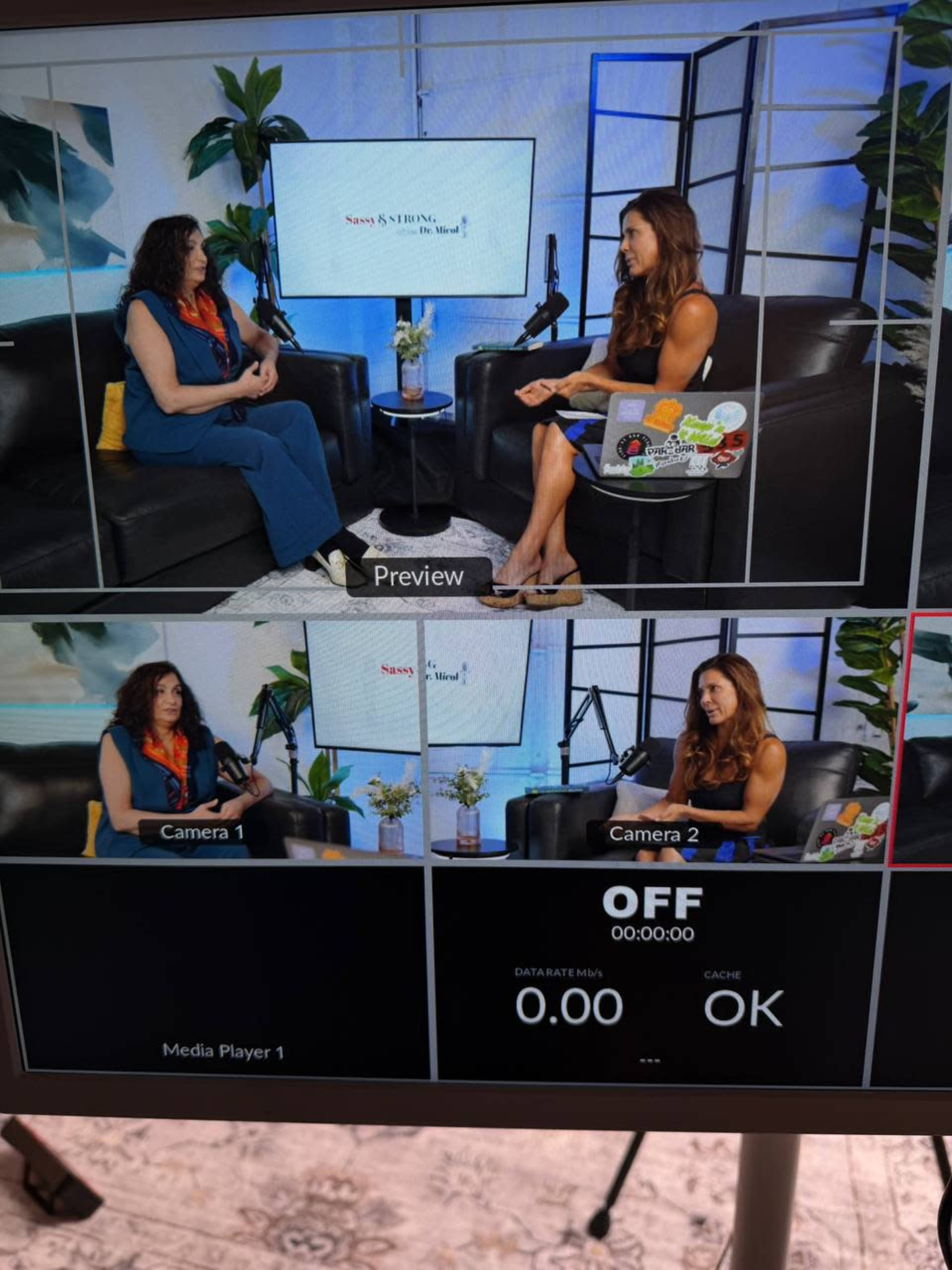 Two women are seated in a studio setup, engaged in conversation, with one woman on camera and the other in the preview screen, surrounded by potted plants and microphones.