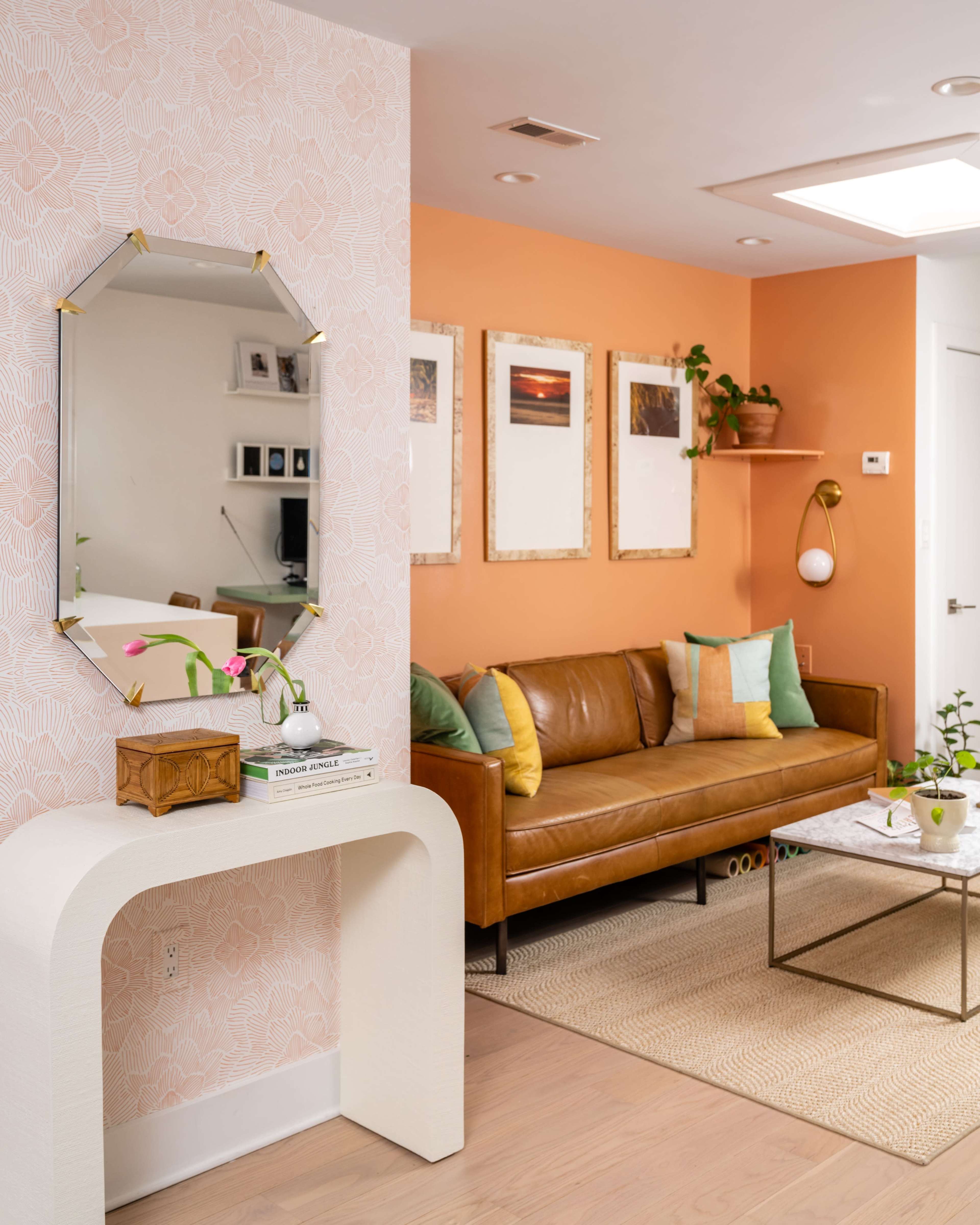 A living room with an orange wall, a brown leather sofa, a decorative mirror, and framed artwork hanging above a coffee table.